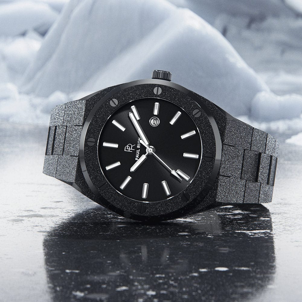Paul Rich frosted black watch with sunray dial and silver markers, placed on icy surface with snow-covered mountains in the background.