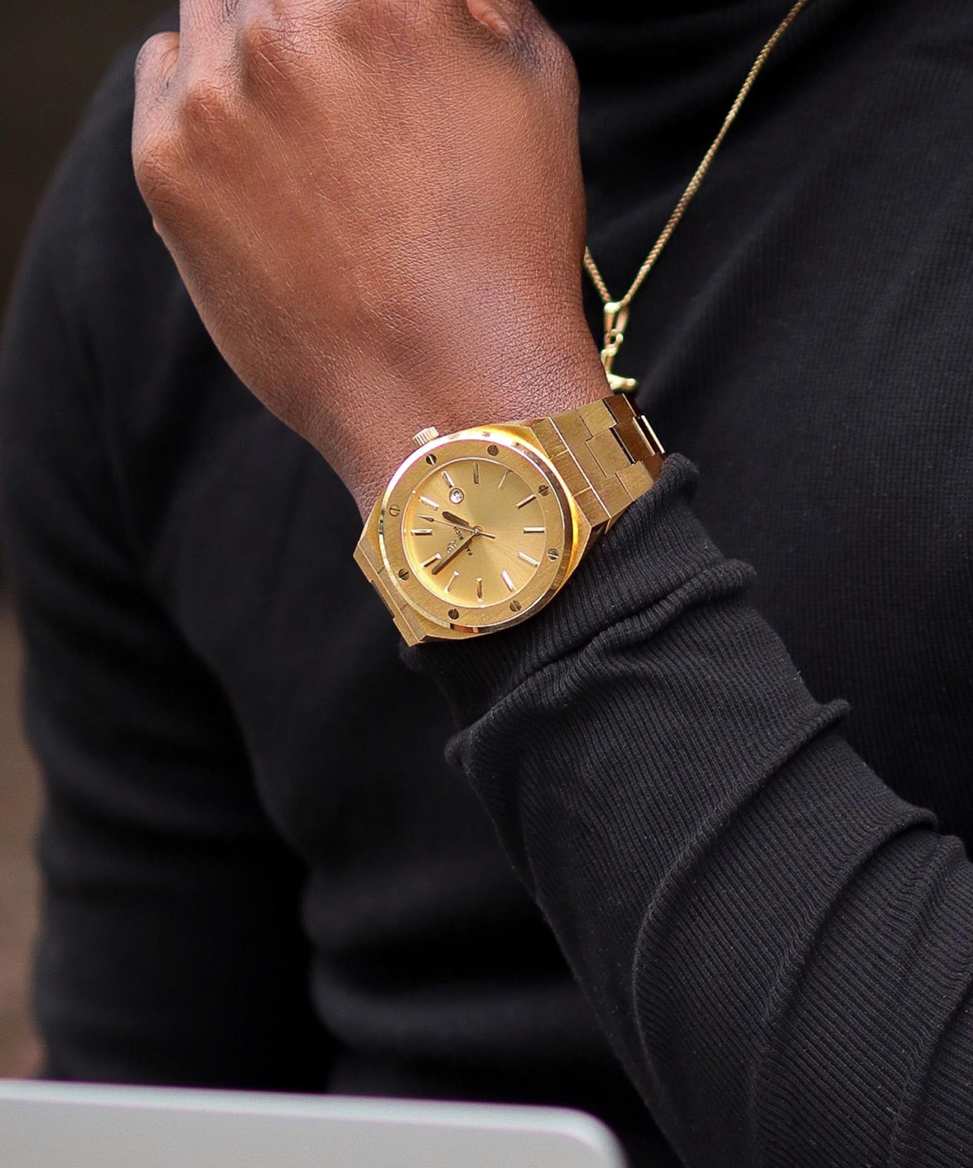 Paul Rich brushed gold watch with gold dial and date window, worn on wrist with black long sleeve shirt and gold necklace.