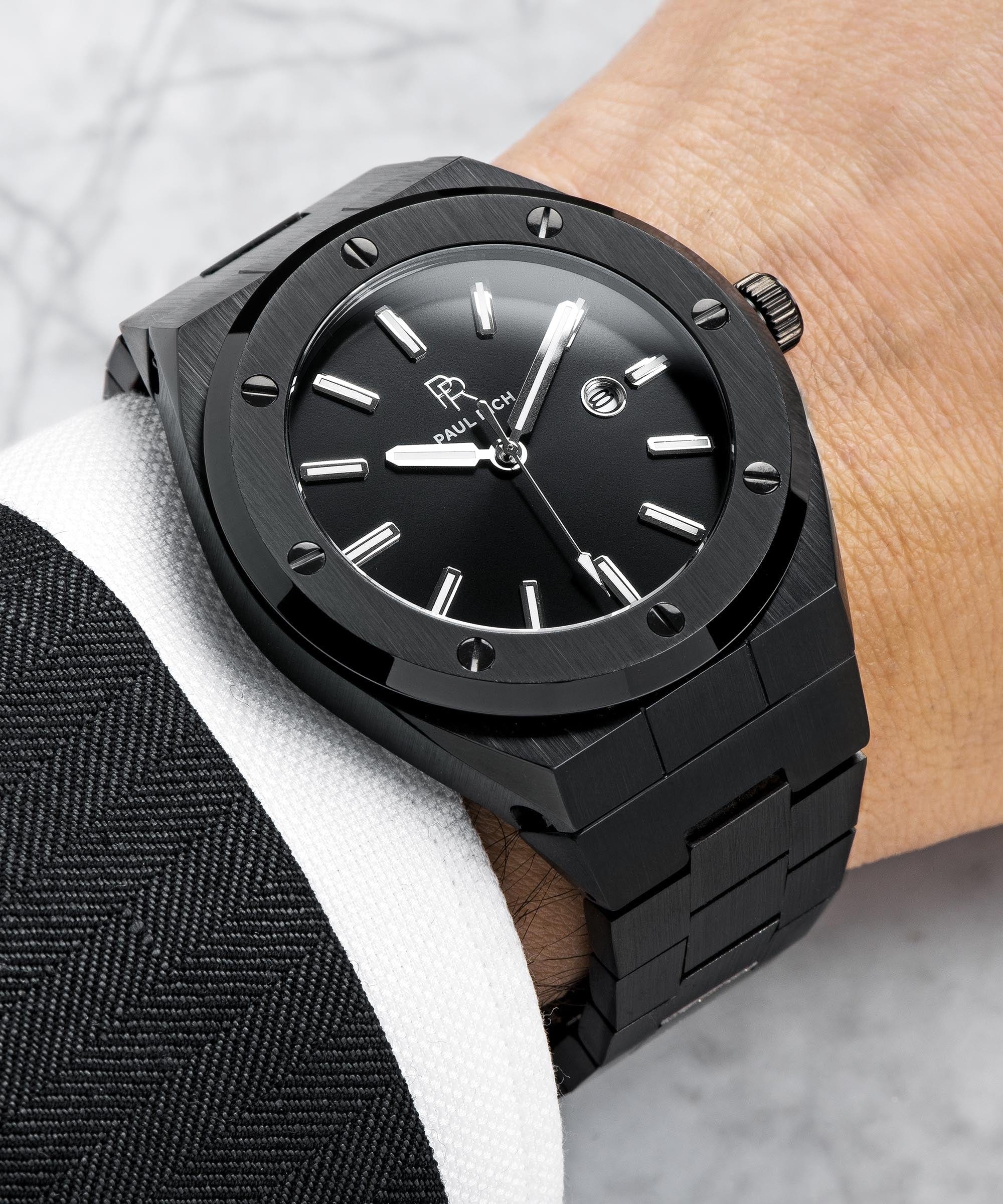 Paul Rich brushed black watch with black sunray dial and date window, worn with black suit jacket and white shirt cuff.