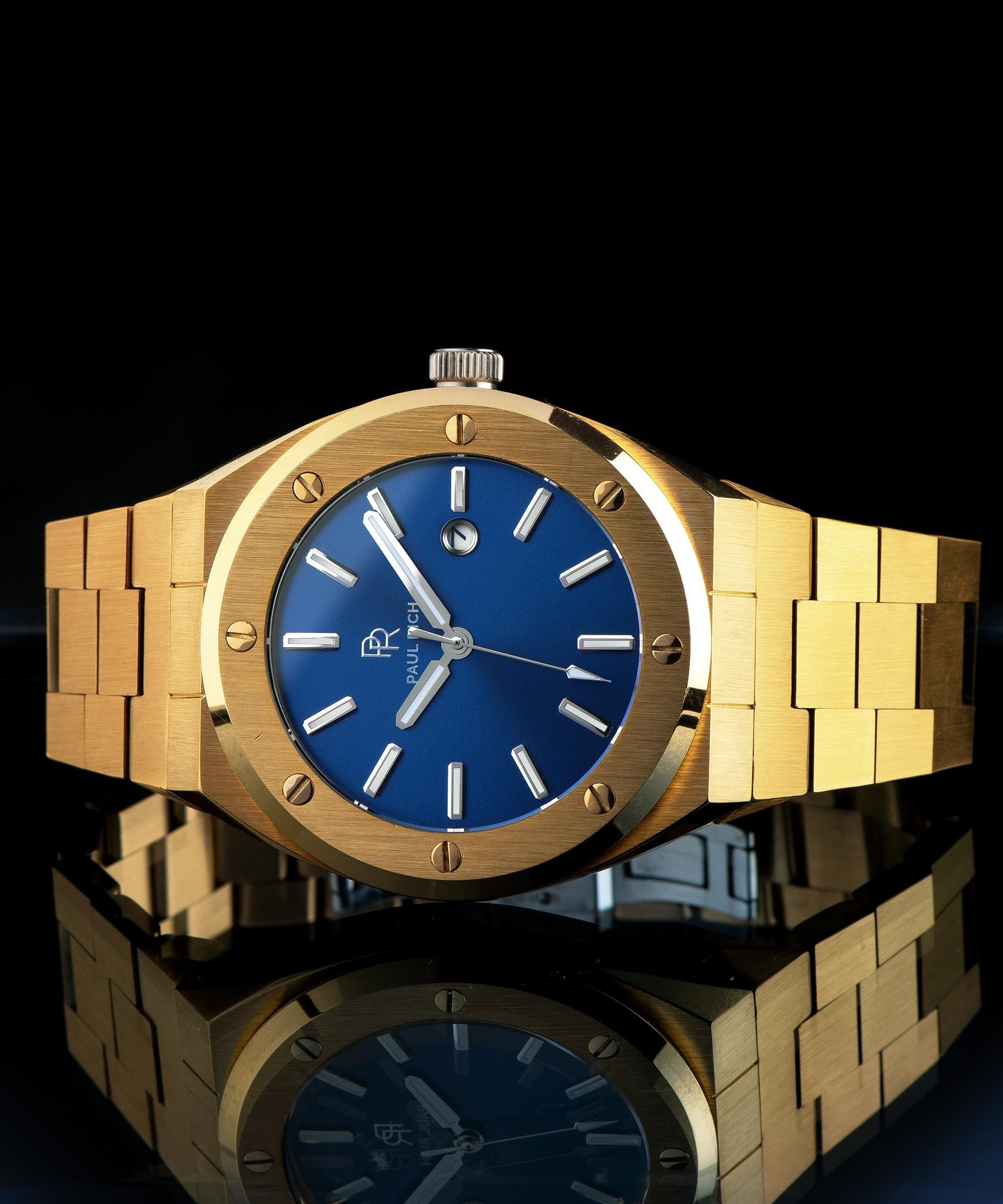 Paul Rich brushed gold watch with matte blue dial and silver markers, displayed horizontally on reflective black surface with crown visible.