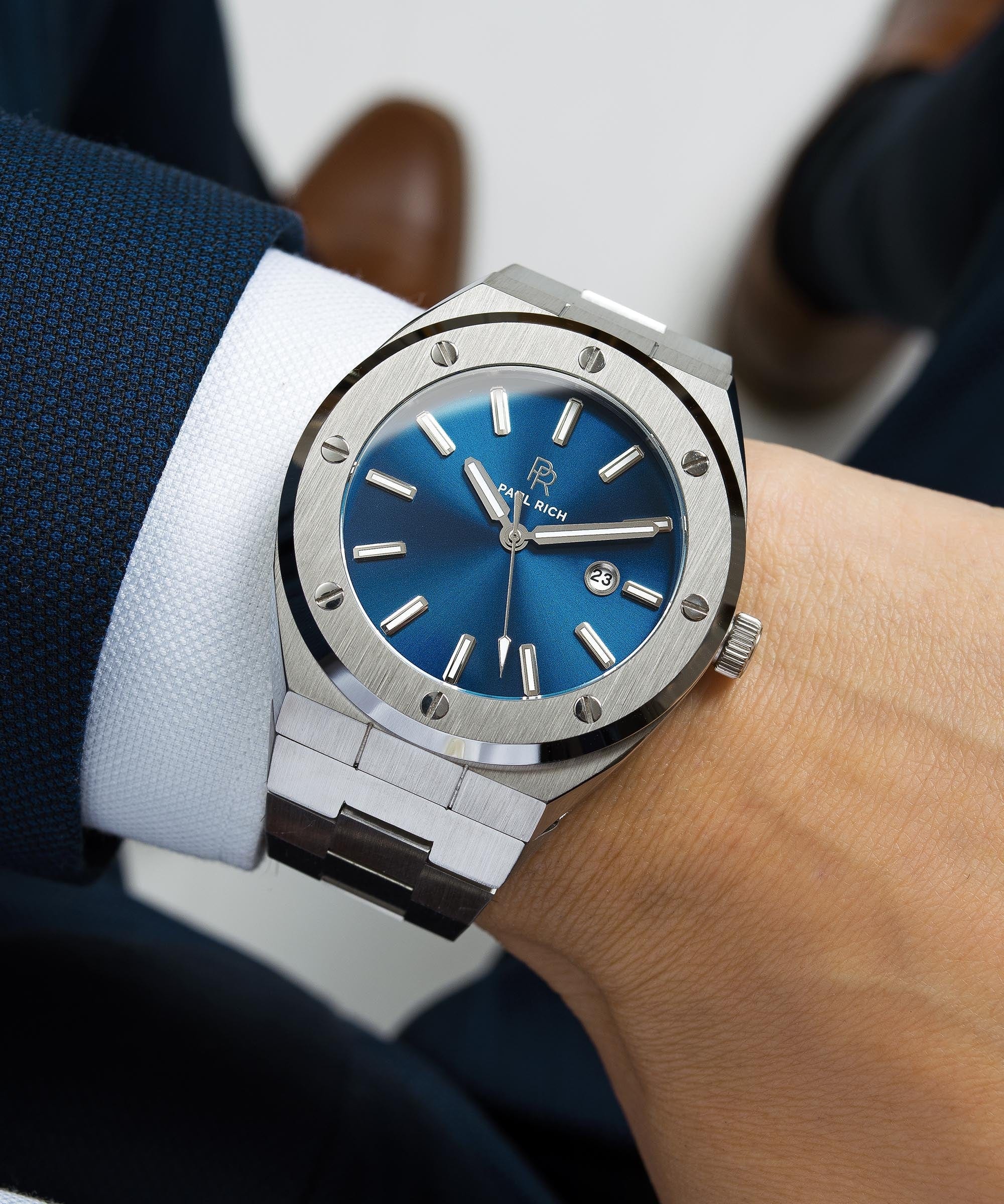 Paul Rich brushed silver watch with deep blue sunray dial and date window, worn with navy suit jacket and white shirt cuff.