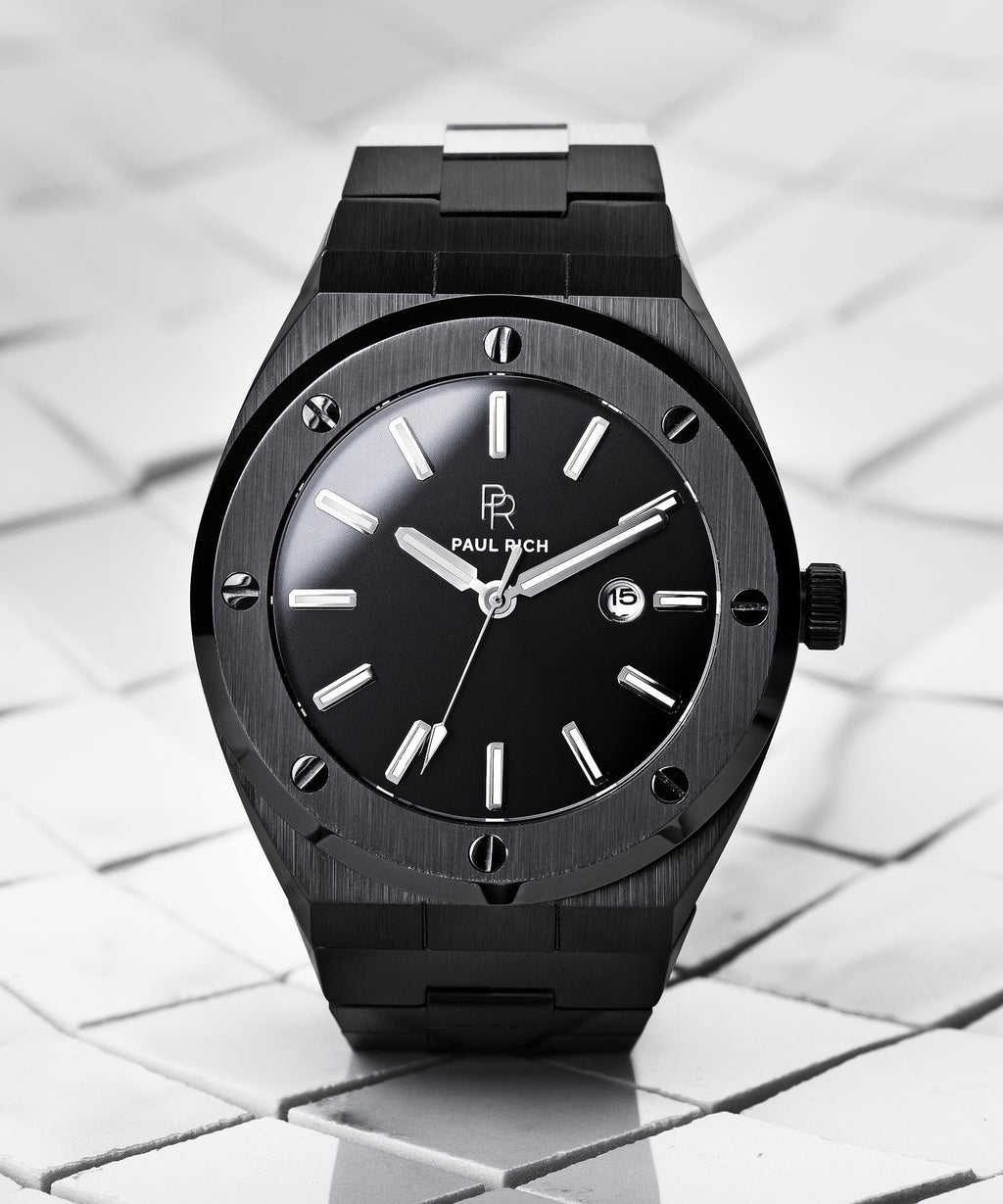 Paul Rich brushed black watch with black sunray dial and date window, displayed upright on white tile surface with soft shadows.