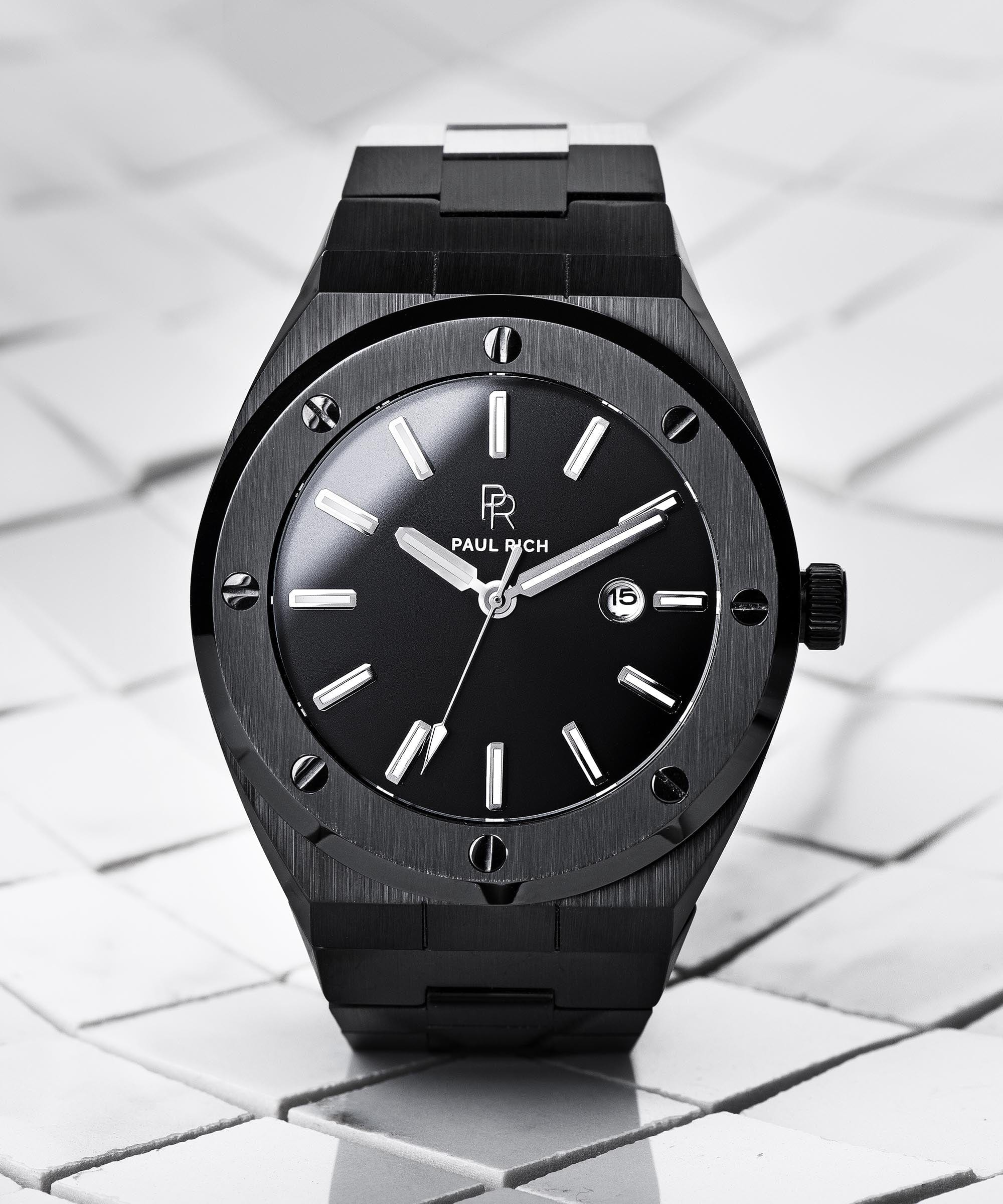 Paul Rich brushed black watch with black sunray dial and date window, displayed upright on white tile surface with soft shadows.