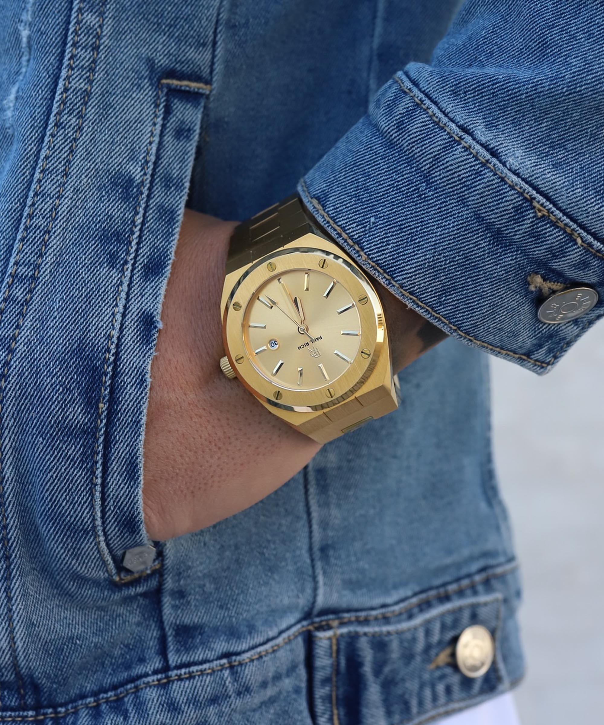 Paul Rich brushed gold watch with gold dial and date window, worn on wrist with blue denim jacket, hand in pocket.