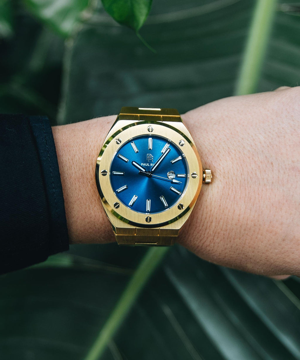 Paul Rich brushed gold watch with deep blue sunray dial and date window, worn on wrist against green tropical leaves in background.