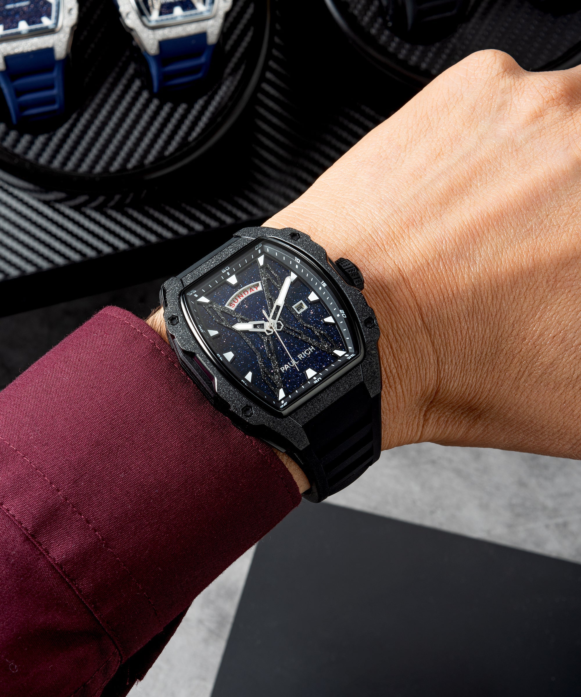 Paul Rich watch with blue aventurine dial and frosted bezel worn on wrist with burgundy shirt; display tray with watches in background.