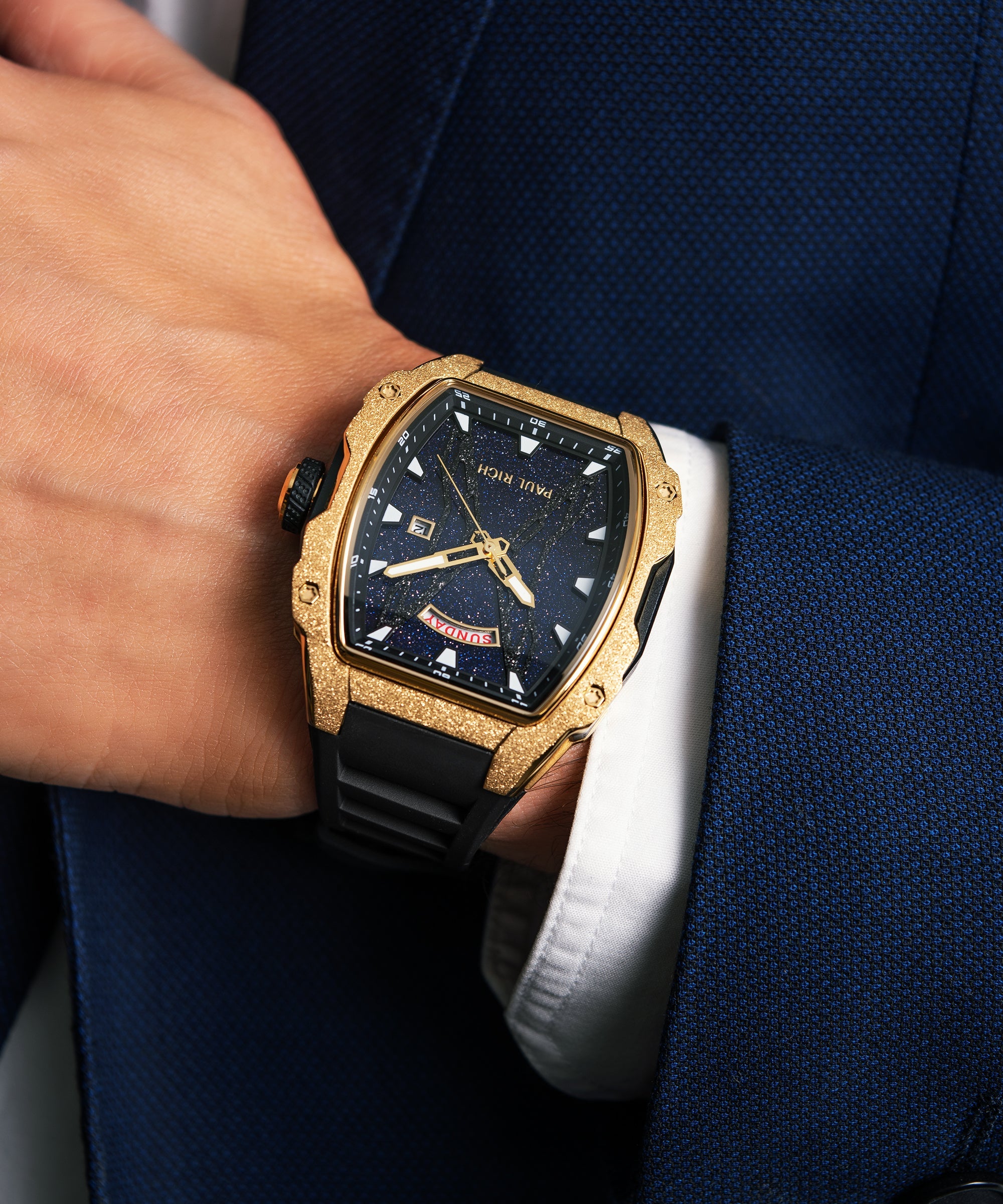 Man wearing a Paul Rich watch with frosted gold tonneau case, blue aventurine dial, and black strap under a blue textured suit sleeve.