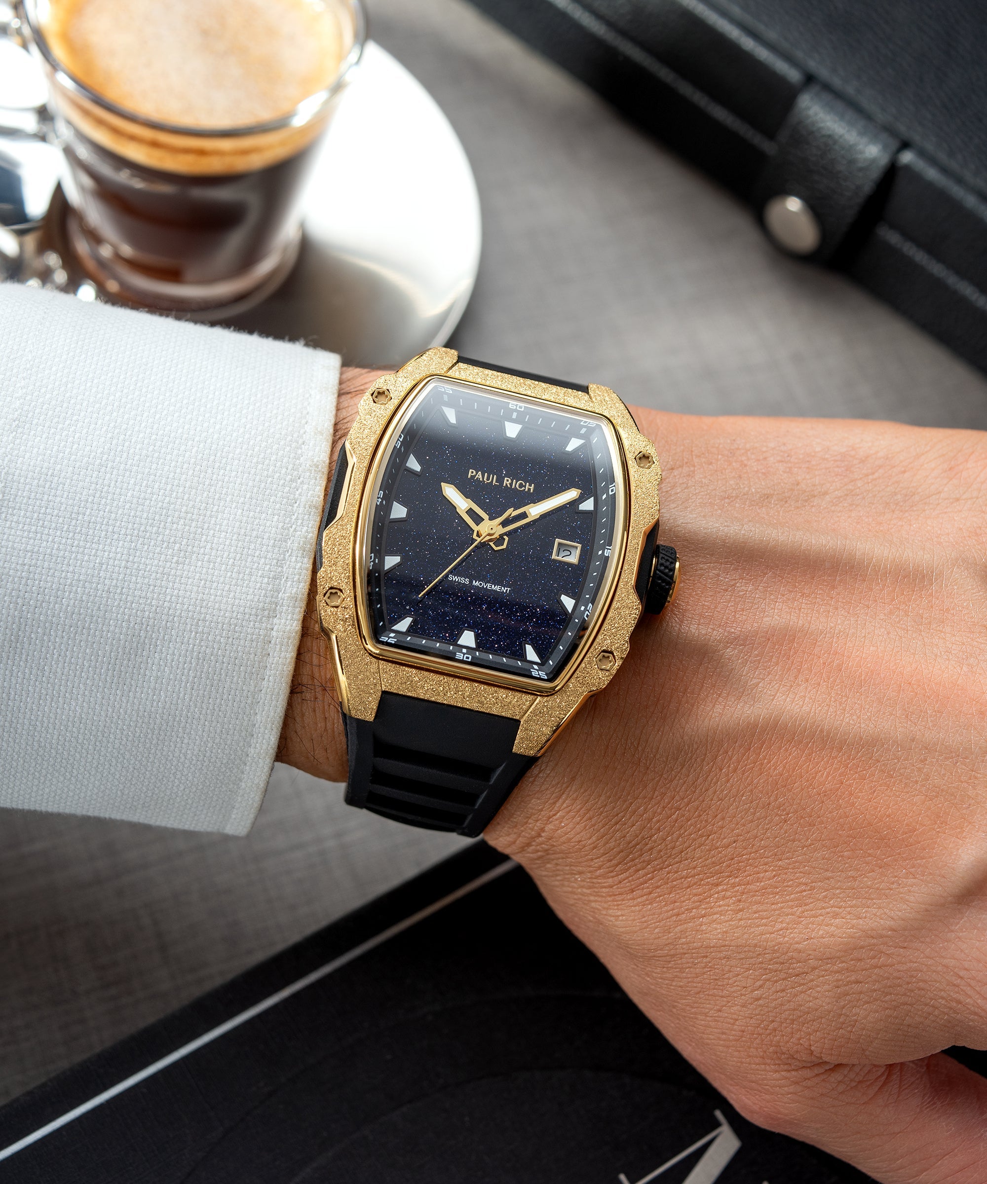 Tonneau-shaped Paul Rich watch with blue aventurine dial and gold frosted bezel worn on wrist with white cuff, coffee in background.