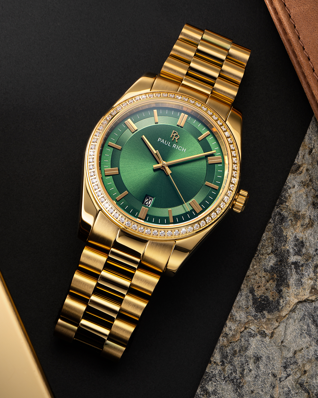 Gold-tone Paul Rich watch with green sunray dial, diamond-set bezel, and gold link bracelet, displayed on a flat lay surface with leather, stone, and matte black textures.