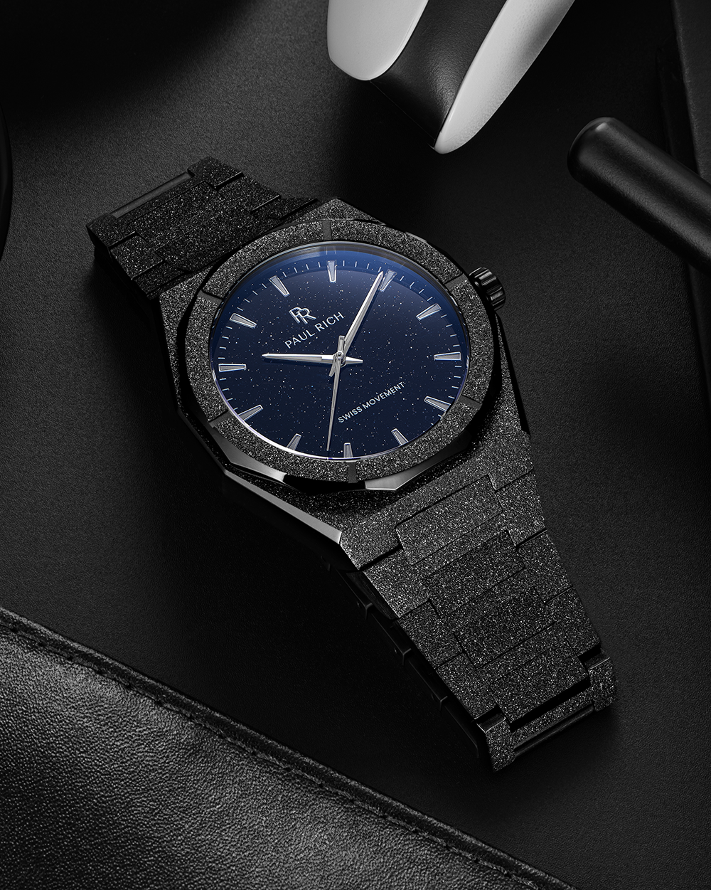 Black frosted Paul Rich watch with blue aventurine dial resting on a black leather surface near gaming gear and a headset.