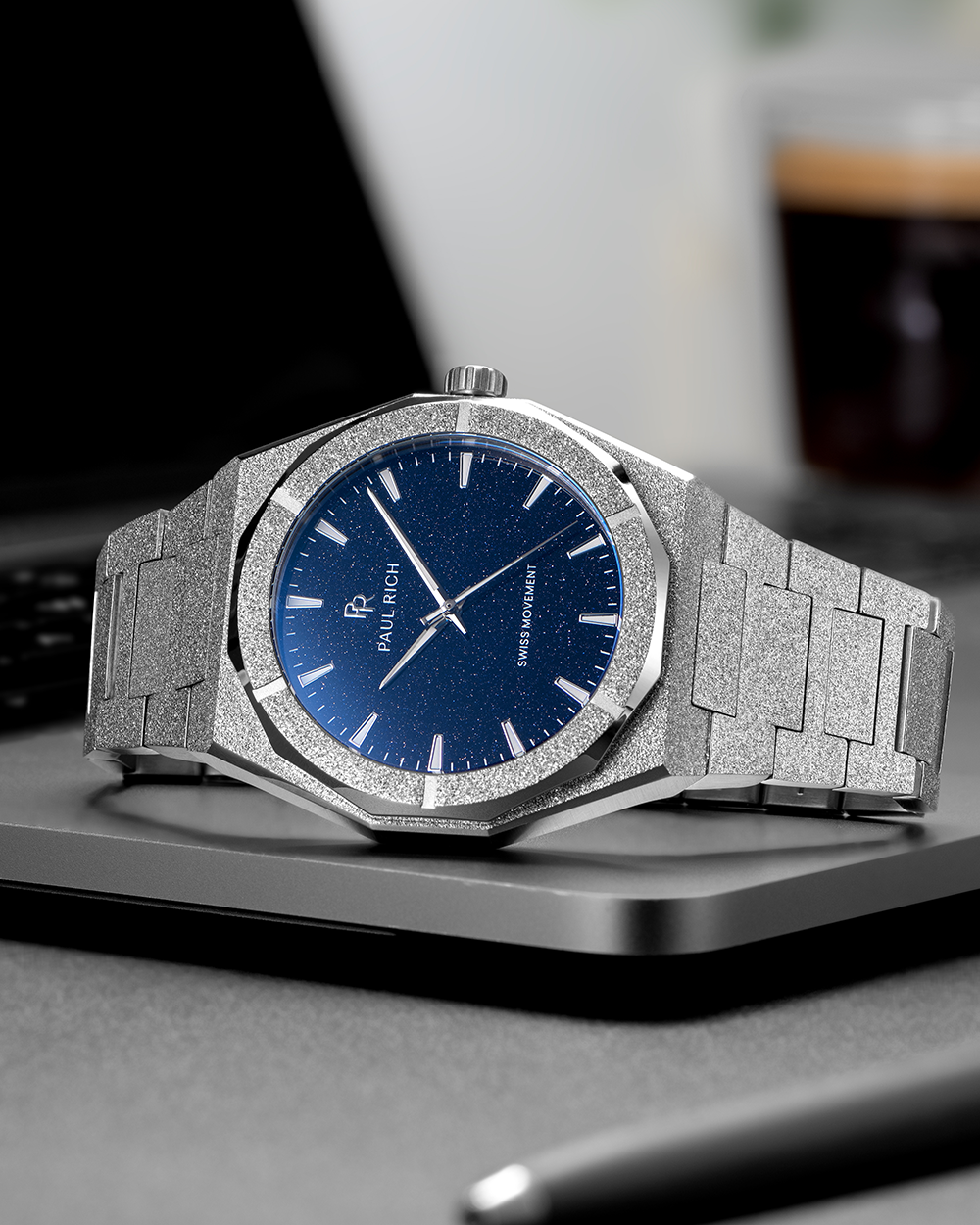 Silver frosted Paul Rich watch with blue aventurine dial resting on a closed laptop, with a blurred coffee cup in the background.