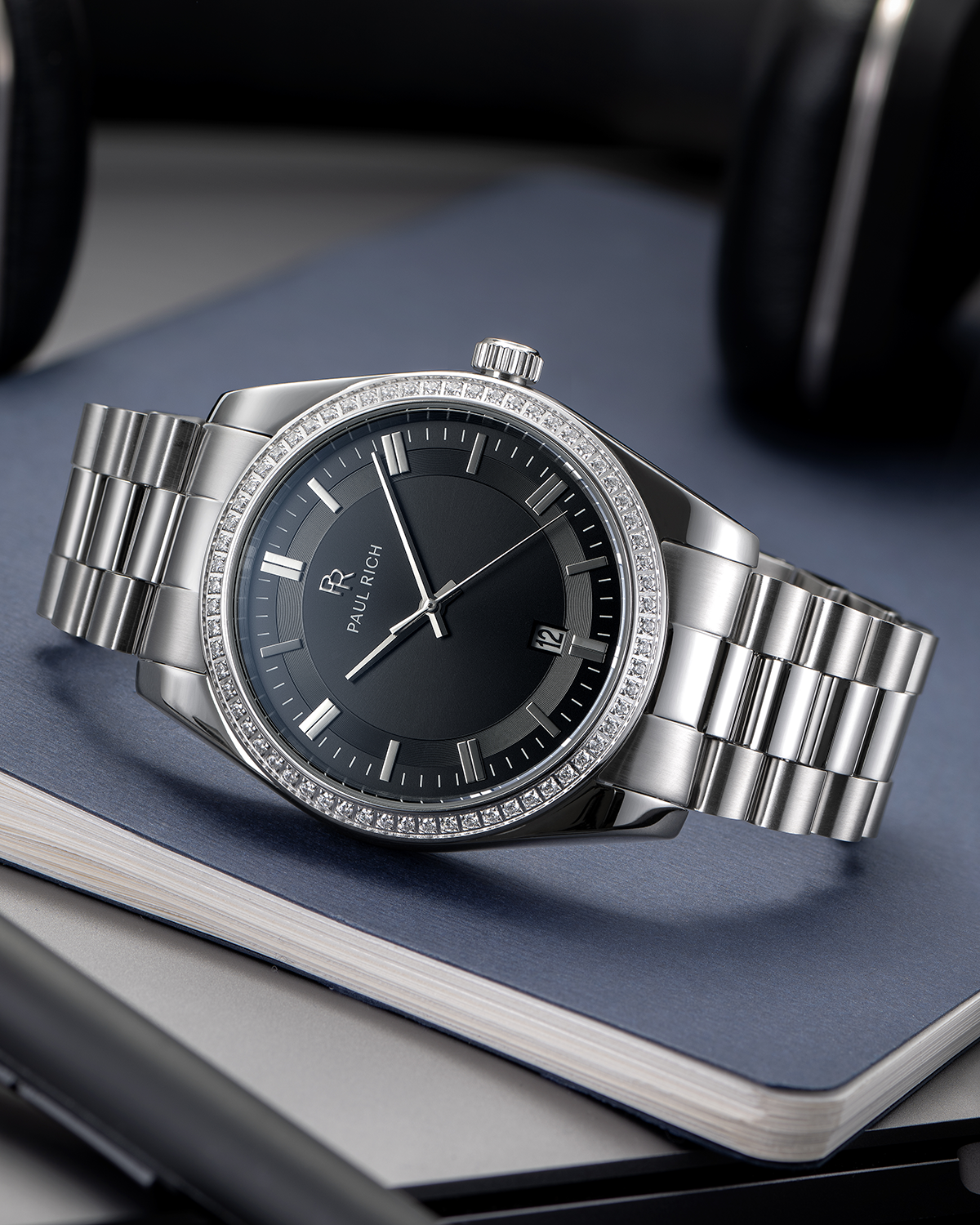 Silver-tone Paul Rich watch with black sunray dial, diamond-set bezel, and silver link bracelet, displayed on top of stacked hardcover notebooks with a pen and headphones nearby.