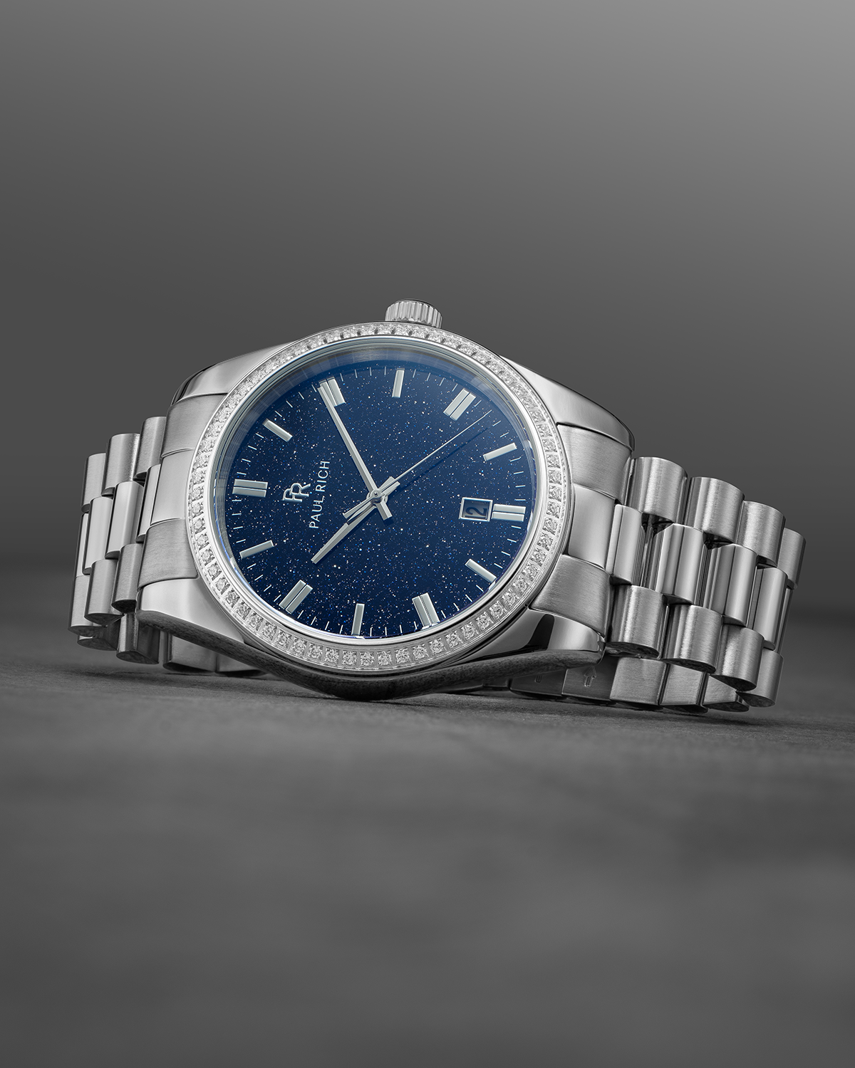 Silver wristwatch with a blue speckled dial, crystal-set bezel, and metal bracelet displayed on a dark gray surface with a gradient background.