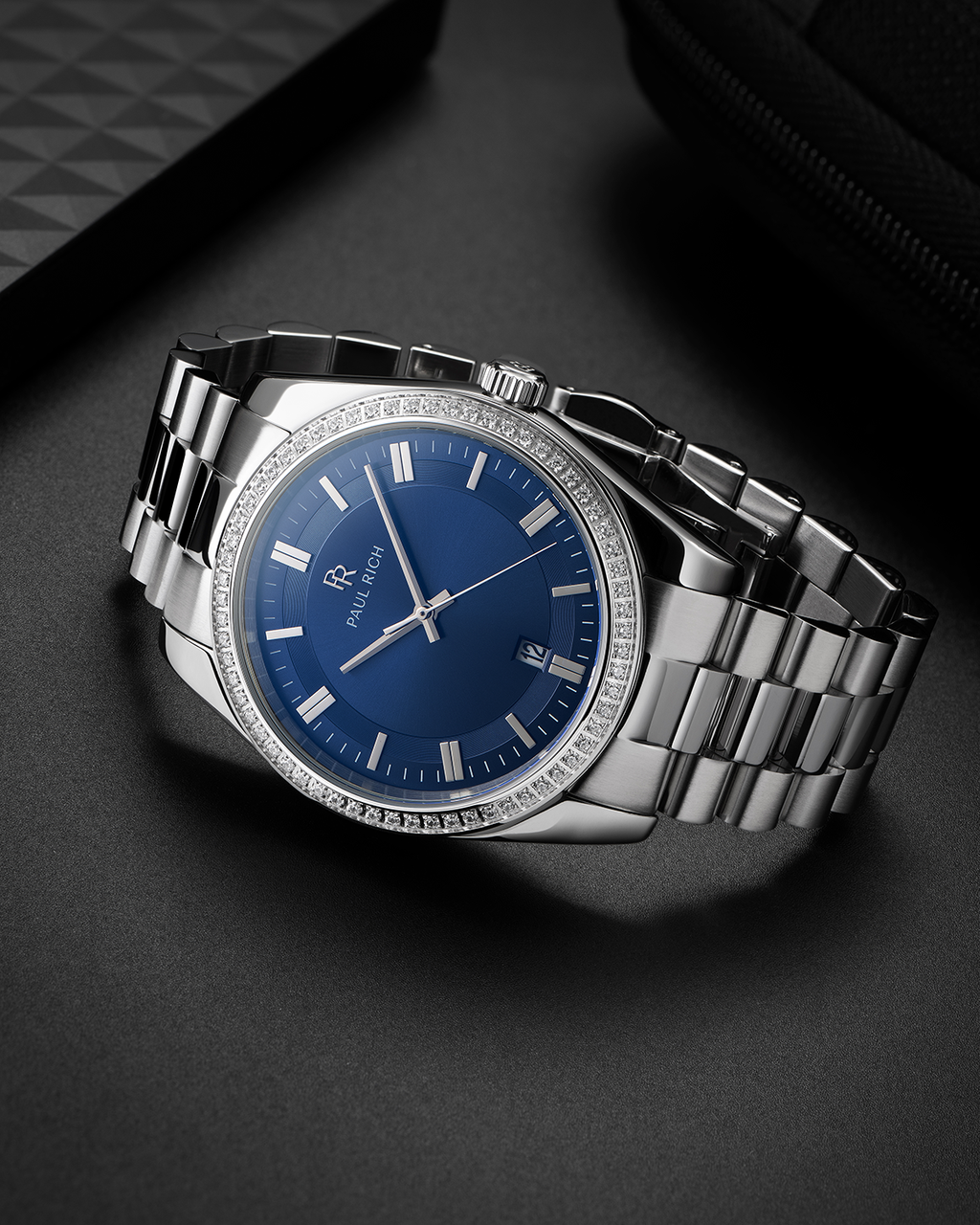 Silver-tone Paul Rich watch with blue sunray dial, diamond-set bezel, and silver link bracelet, displayed on a dark matte surface beside textured black accessories.