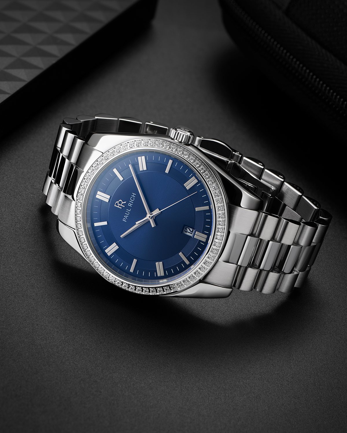 Silver-tone Paul Rich watch with blue sunray dial, diamond-set bezel, and silver link bracelet, displayed on a dark matte surface beside textured black accessories.