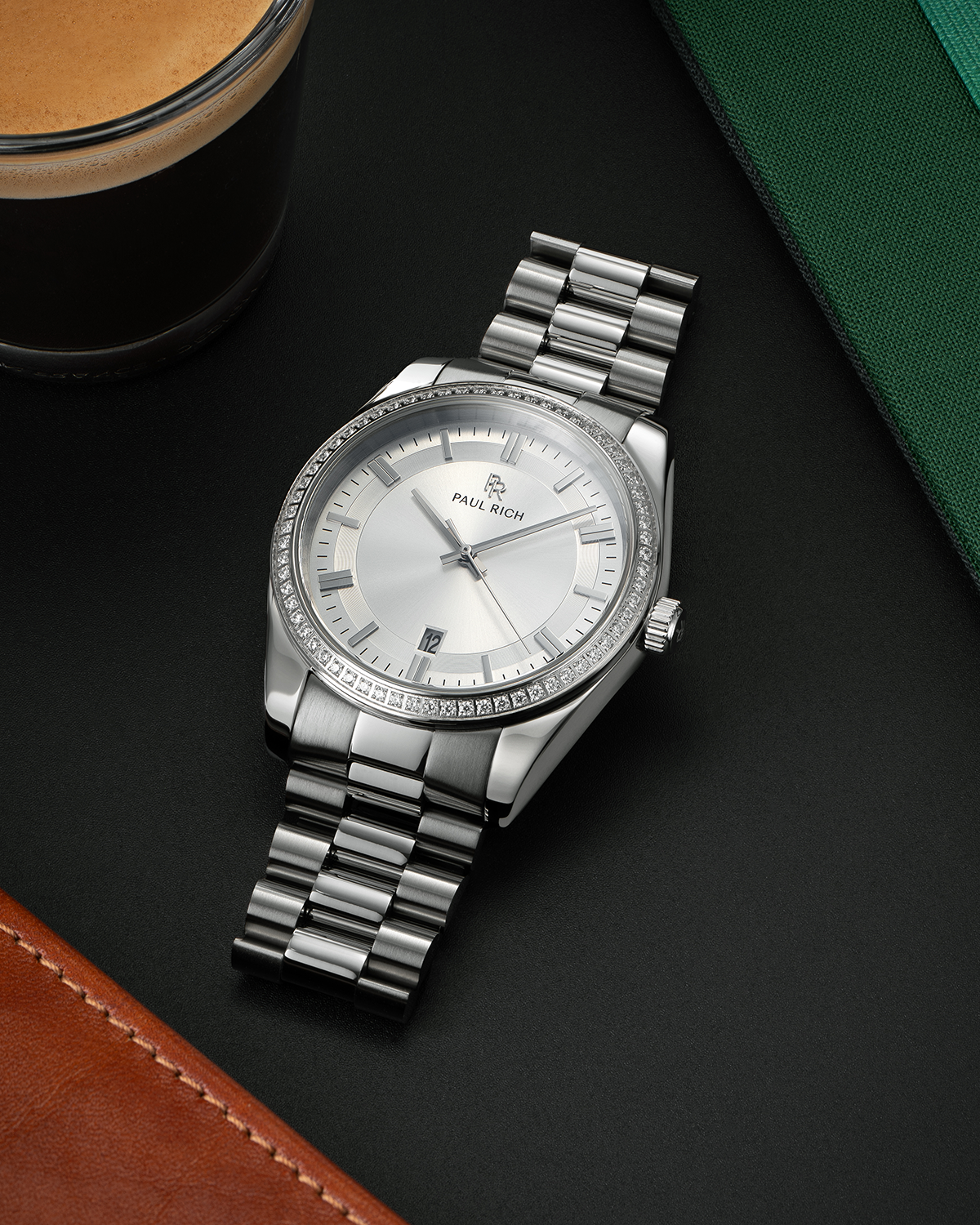 Silver-tone Paul Rich watch with silver sunray dial, diamond-set bezel, and polished silver link bracelet, displayed on a black surface with a cup of espresso, green notebook, and brown leather item.