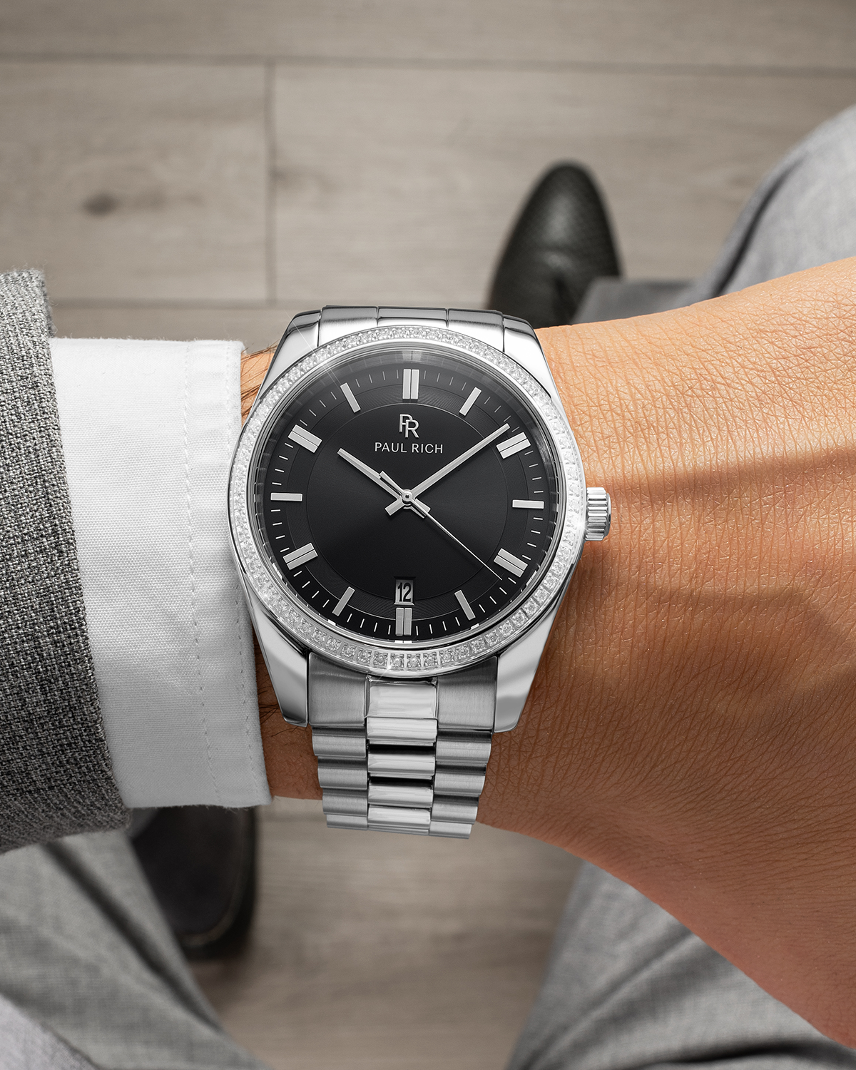 Silver-tone Paul Rich watch with black sunray dial, diamond-set bezel, and silver link bracelet, worn on wrist with a white shirt and grey suit, shown from a top-down view above a wooden floor.