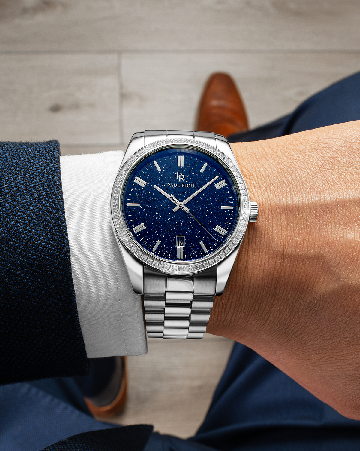 Close-up of a silver wristwatch with a blue speckled dial, worn on a man's wrist in a navy suit and brown shoes, sitting indoors.