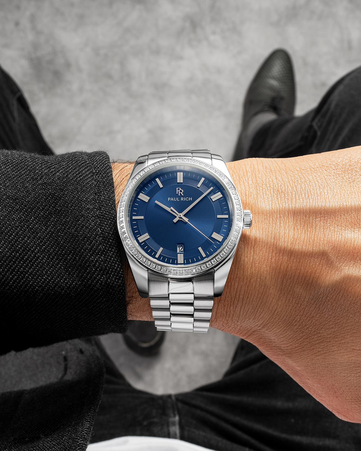 Silver-tone Paul Rich watch with blue sunray dial, diamond-set bezel, and silver link bracelet, worn on wrist with a black jacket and black pants, shown from above with gray flooring in the background.