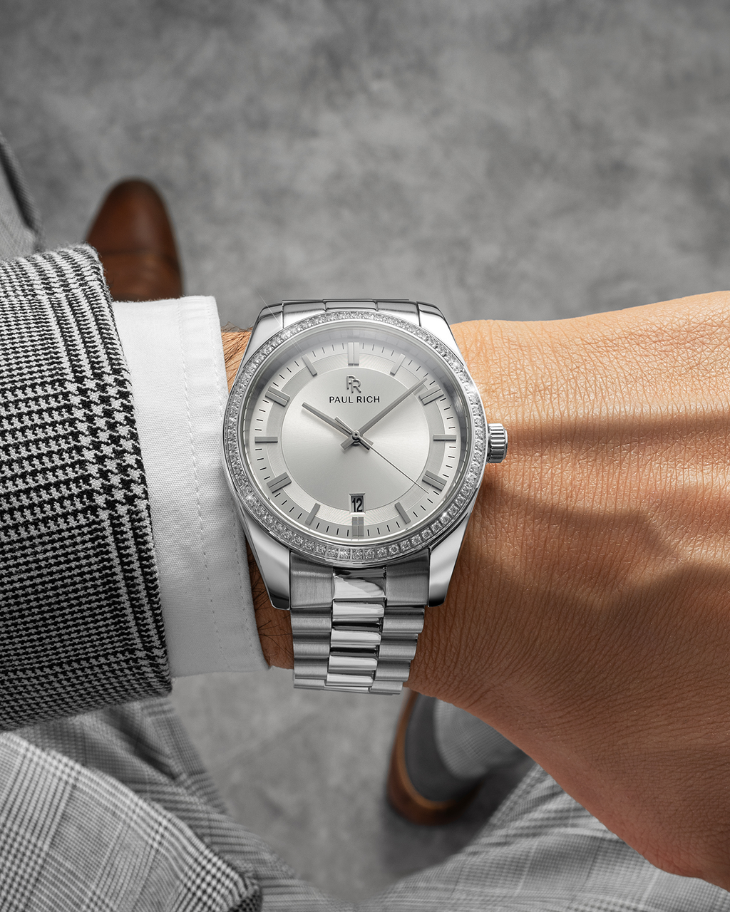 Silver-tone Paul Rich watch with silver sunray dial, diamond-set bezel, and silver link bracelet, worn on wrist with a houndstooth blazer and white cuff, shown from above against a grey floor.