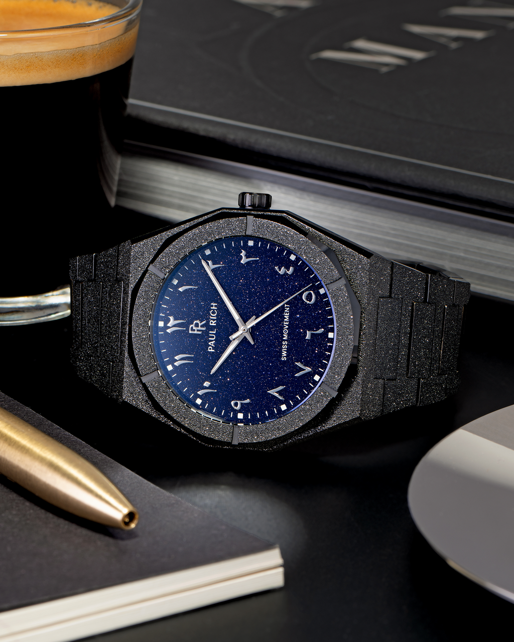 Frosted black watch with a blue aventurine dial and Arabic numerals, placed on a black desk next to a glass of espresso, gold pen, and notebook.