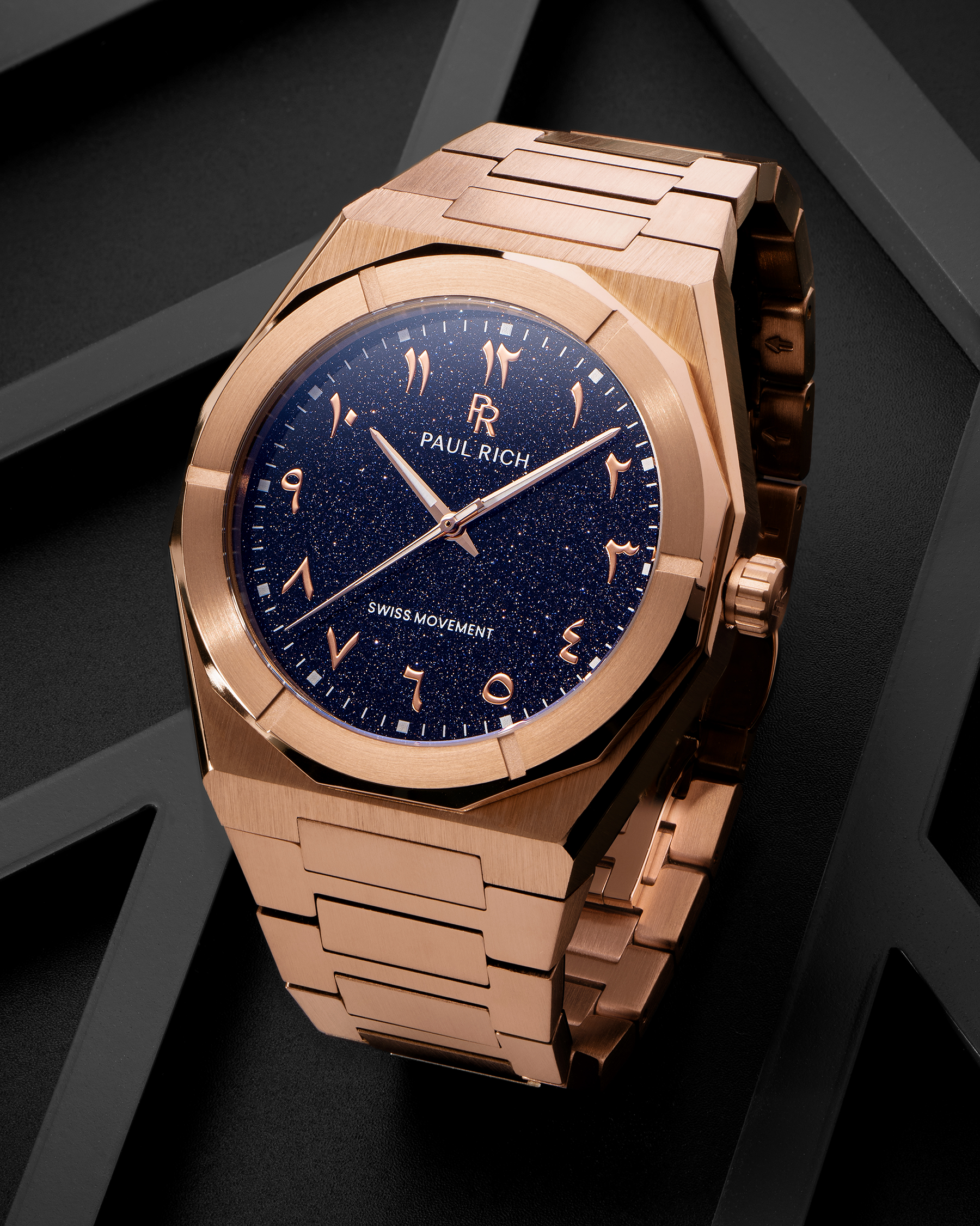 Paul Rich watch with brushed rose gold case and blue aventurine dial with Arabic numerals, displayed on a matte black geometric surface.