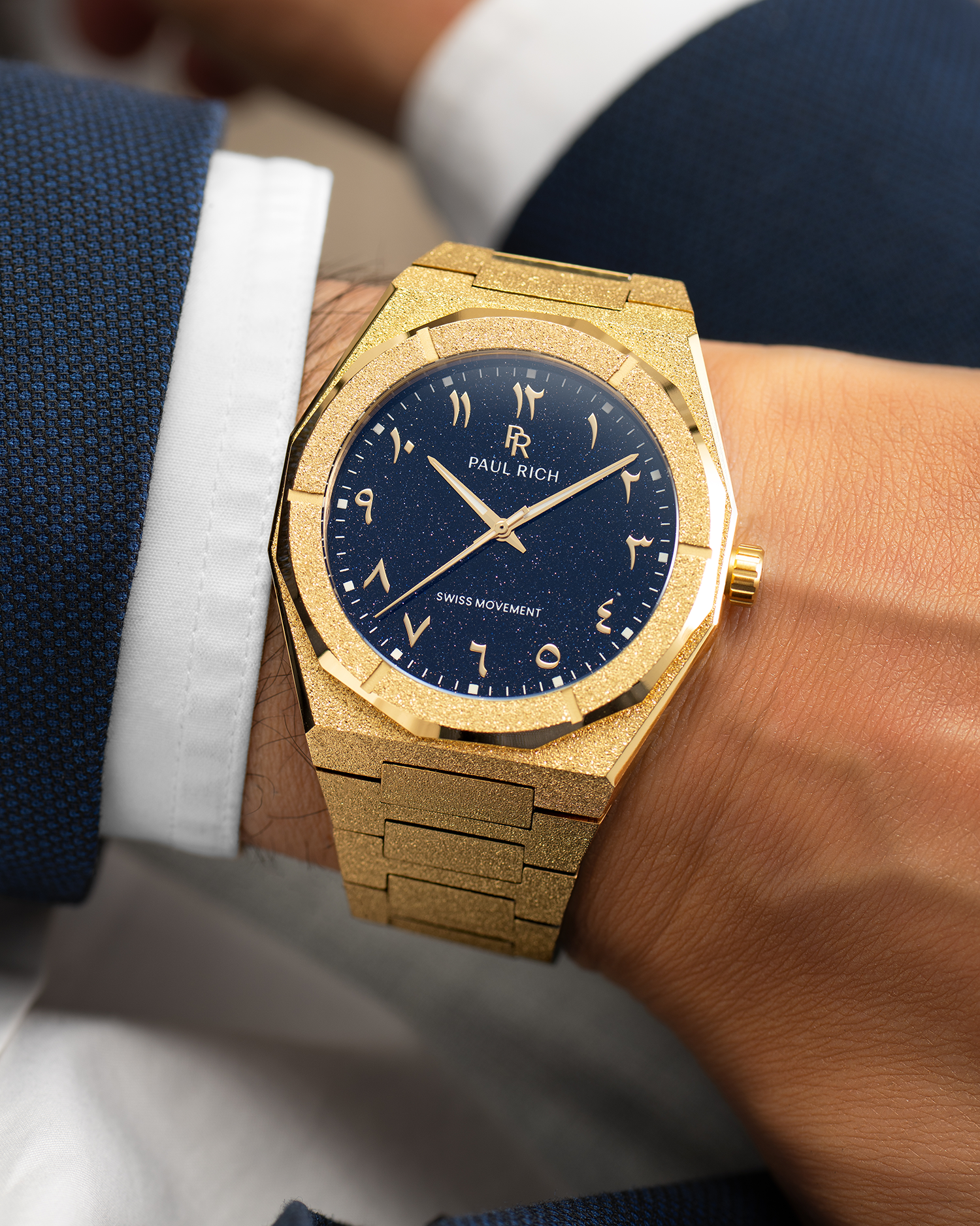 Frosted gold watch with a blue aventurine dial and Arabic numerals, worn with a white cuff and dark blue textured suit.