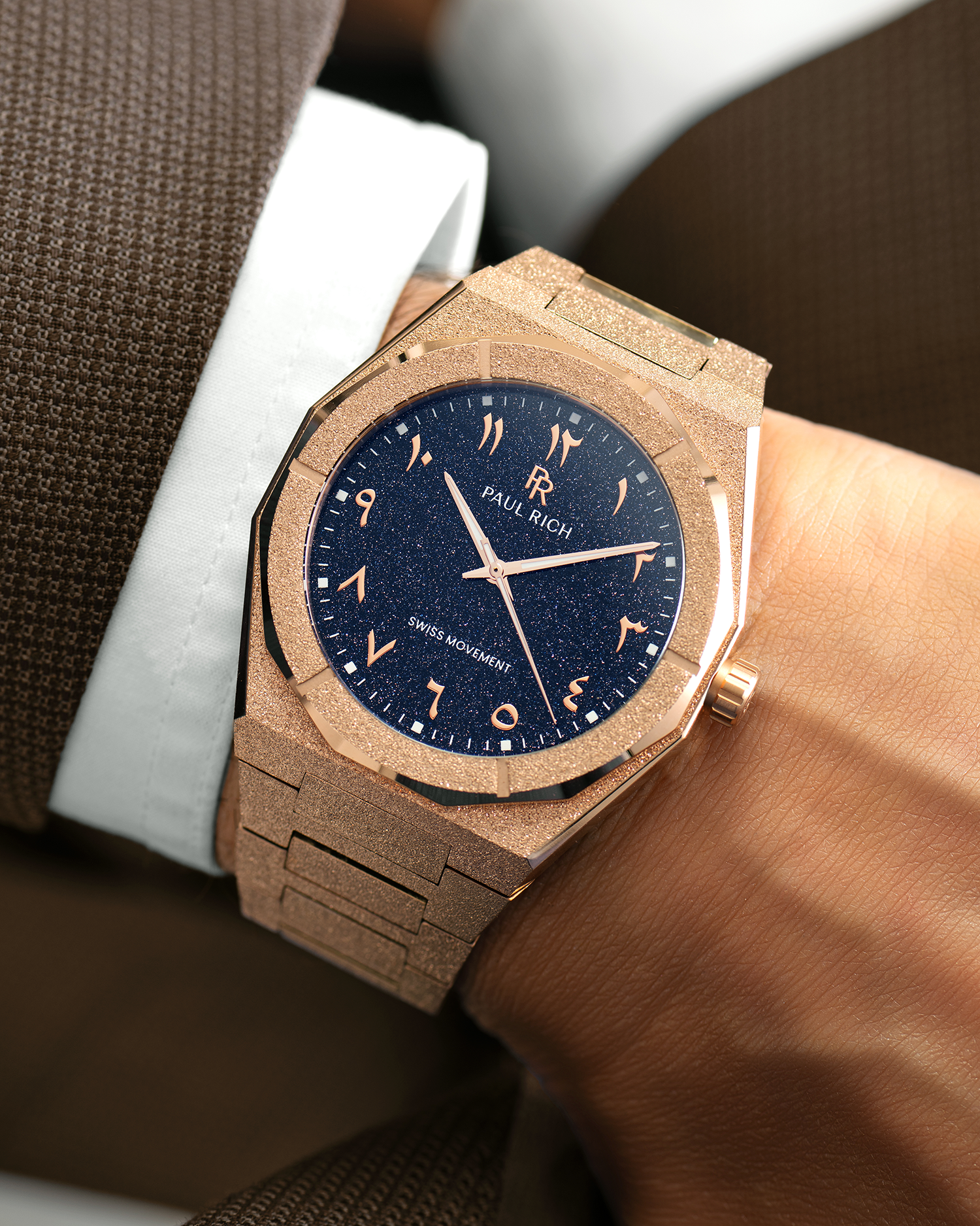 Frosted rose gold watch with blue aventurine dial and Arabic numerals, worn with a white cuff and brown textured suit.