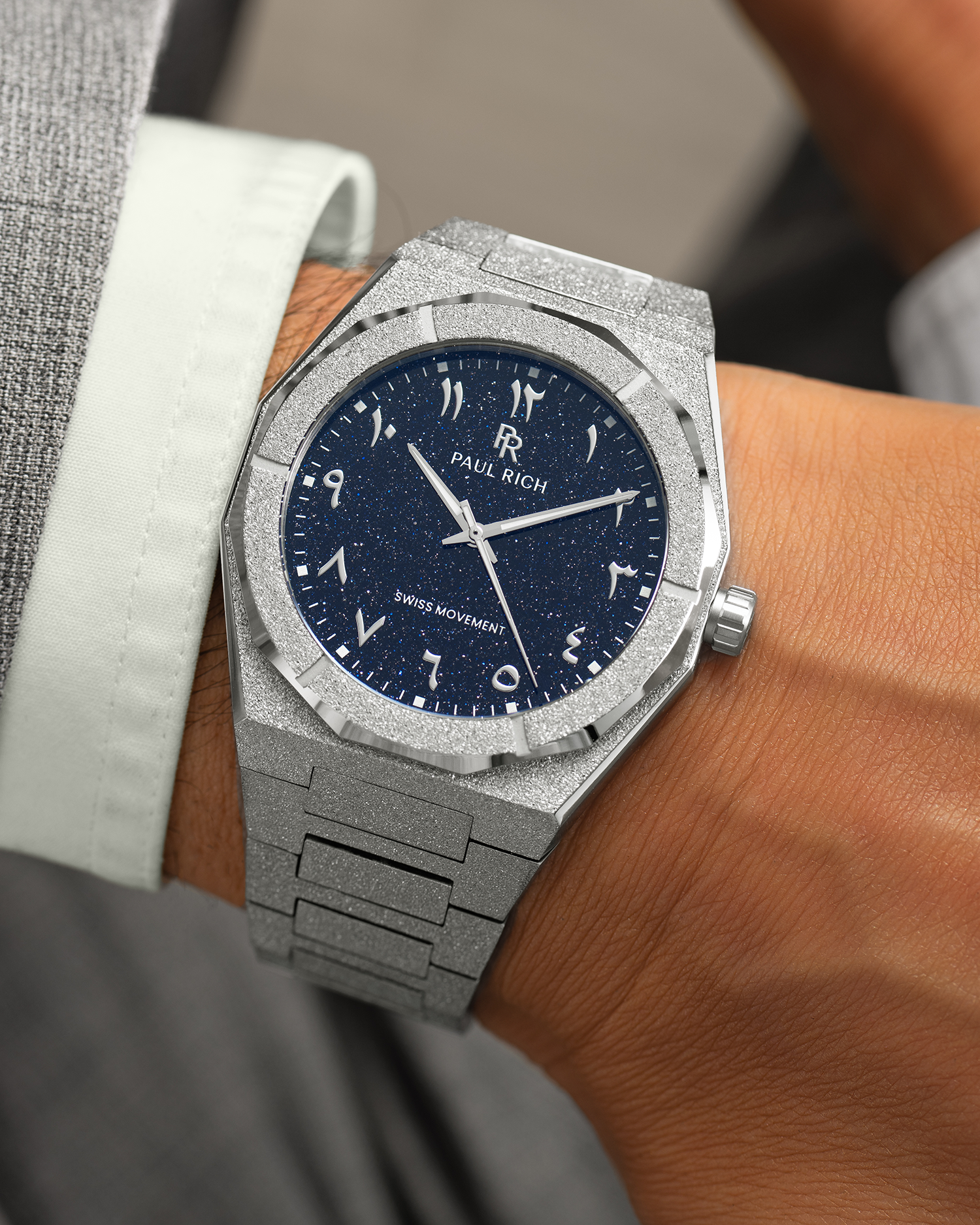 Frosted silver watch with a blue aventurine dial and Arabic numerals, worn with a light green cuff and grey suit.