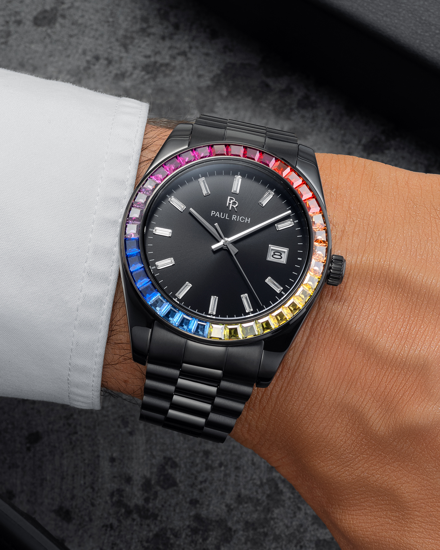 Black wristwatch with a dark dial, baguette hour markers, rainbow bezel, and black metal bracelet worn on a wrist with a white shirt sleeve.