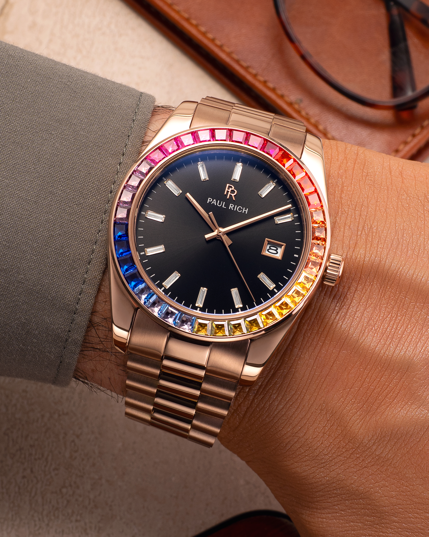 Rose gold wristwatch with a black dial and rainbow bezel worn on a wrist with a green shirt sleeve, next to a brown leather case and glasses.