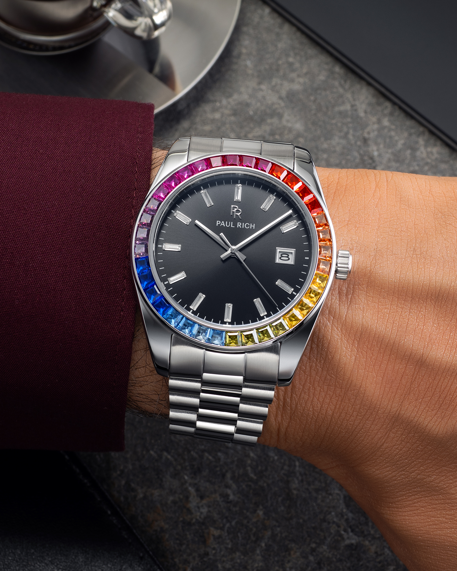 Silver wristwatch with a black dial and rainbow bezel worn on a wrist with a burgundy shirt sleeve, resting on a dark stone surface near a metal cup.