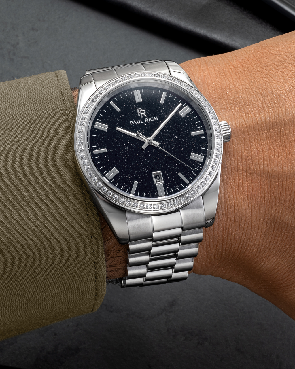 Silver wristwatch with a black aventurine dial, crystal-set bezel, and metal bracelet worn on a wrist with an olive green sleeve on a dark surface.