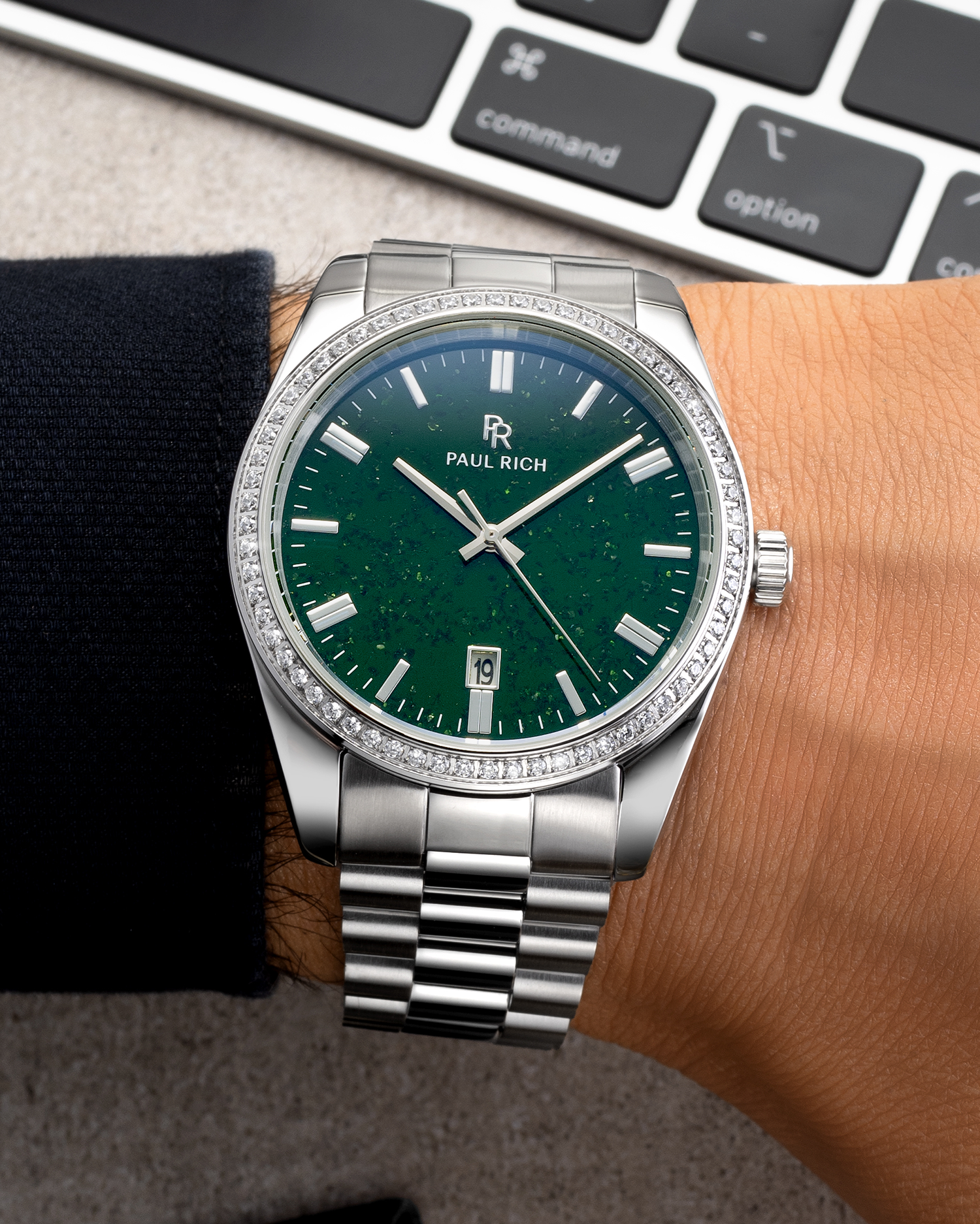 Silver wristwatch with a green aventurine dial, crystal-set bezel, and metal bracelet worn on a wrist near a keyboard on a desk surface.
