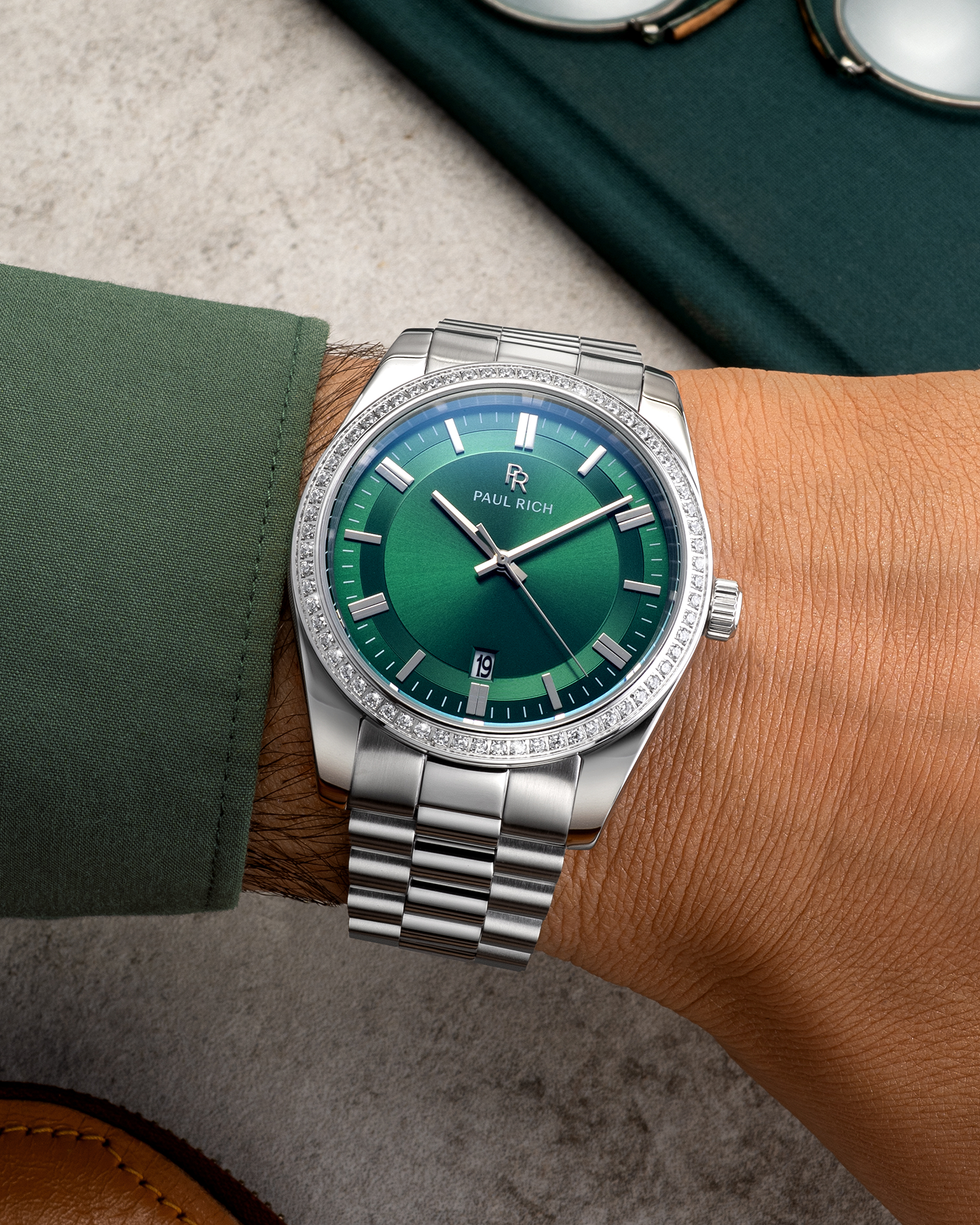 Silver wristwatch with a green sunburst dial, crystal-set bezel, and metal bracelet worn on a wrist with a green shirt sleeve.