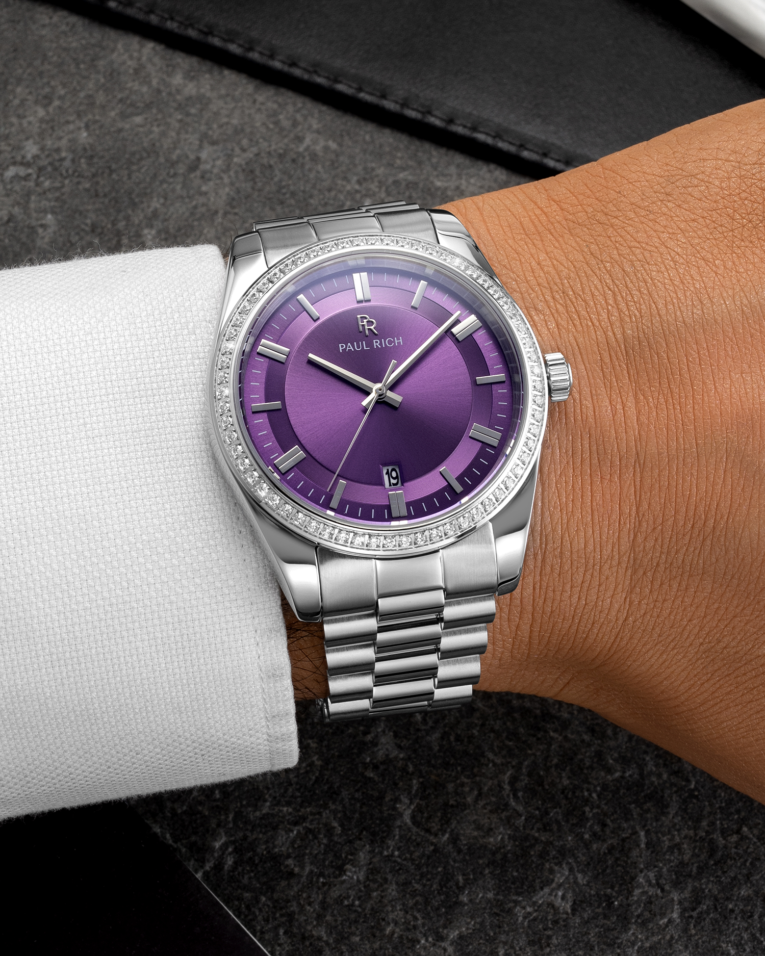 Silver wristwatch with a purple sunburst dial, crystal-set bezel, and metal bracelet worn on a wrist with a white shirt sleeve over a dark surface.