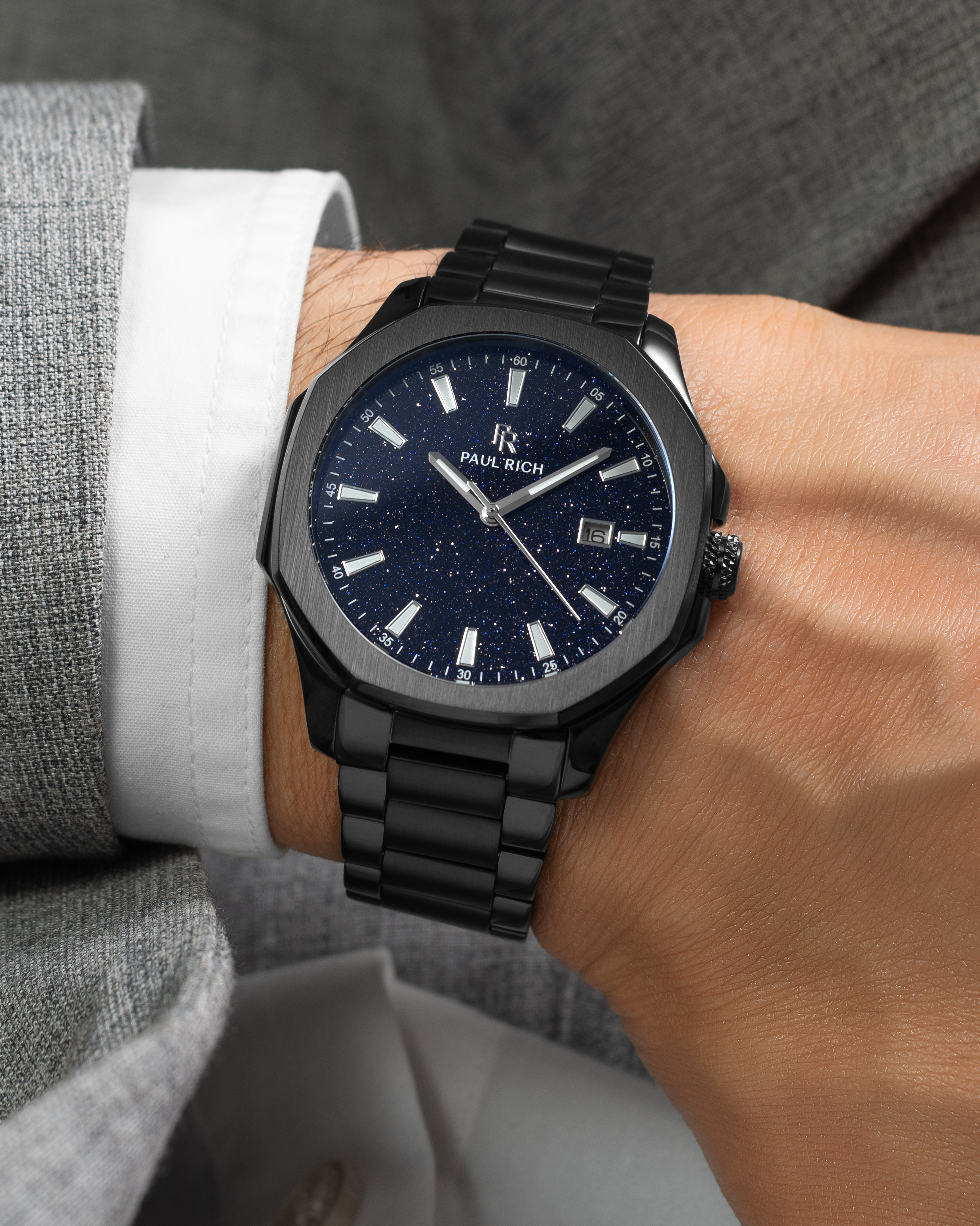 Black Paul Rich watch with blue aventurine dial and date window, worn on wrist with white cuff and light grey textured suit.