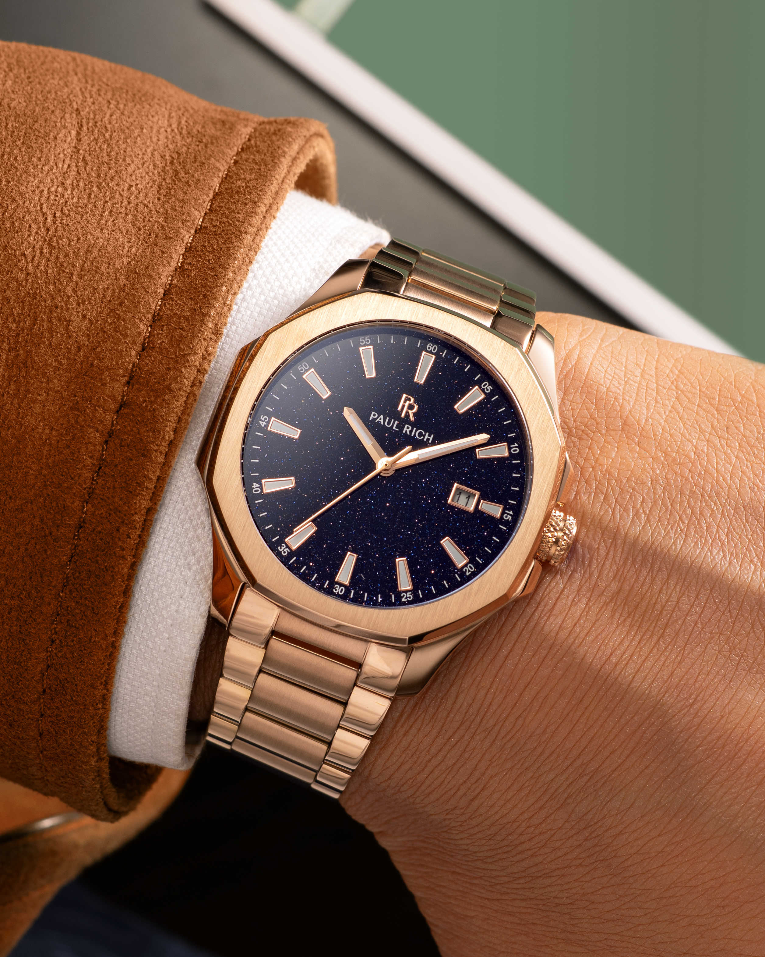 Rose gold Paul Rich watch with blue aventurine dial and date window, worn on wrist with white cuff and brown suede jacket.