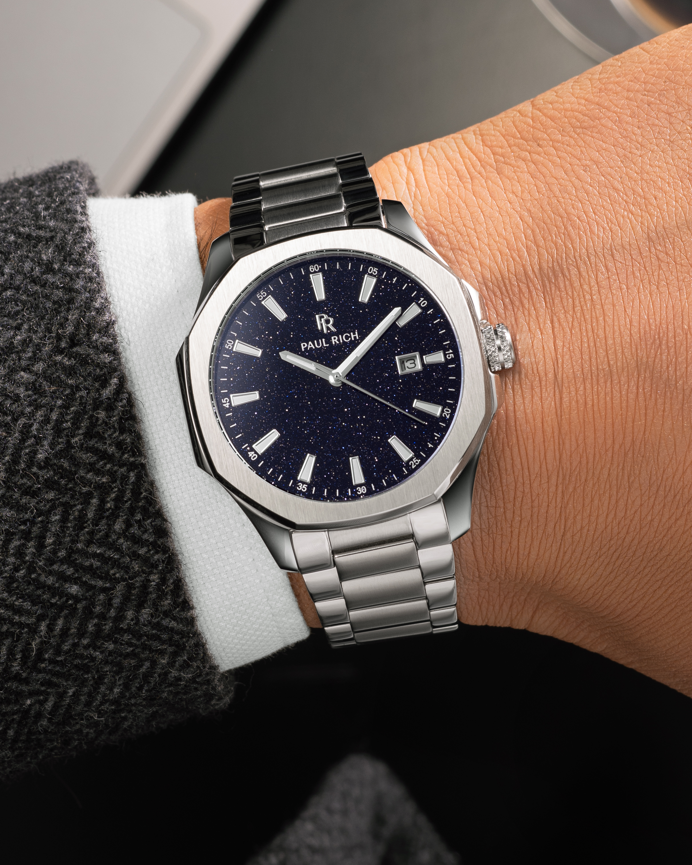 Silver Paul Rich watch with blue aventurine dial and date window, worn on wrist with white shirt and dark herringbone-patterned coat.