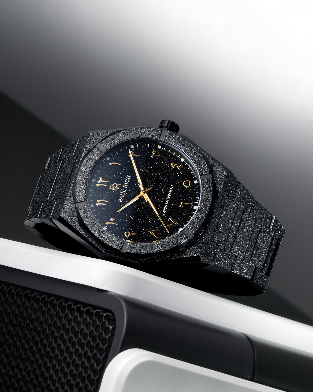 Paul Rich frosted black watch with black aventurine dial and gold Arabic numerals, resting on a silver and black speaker.