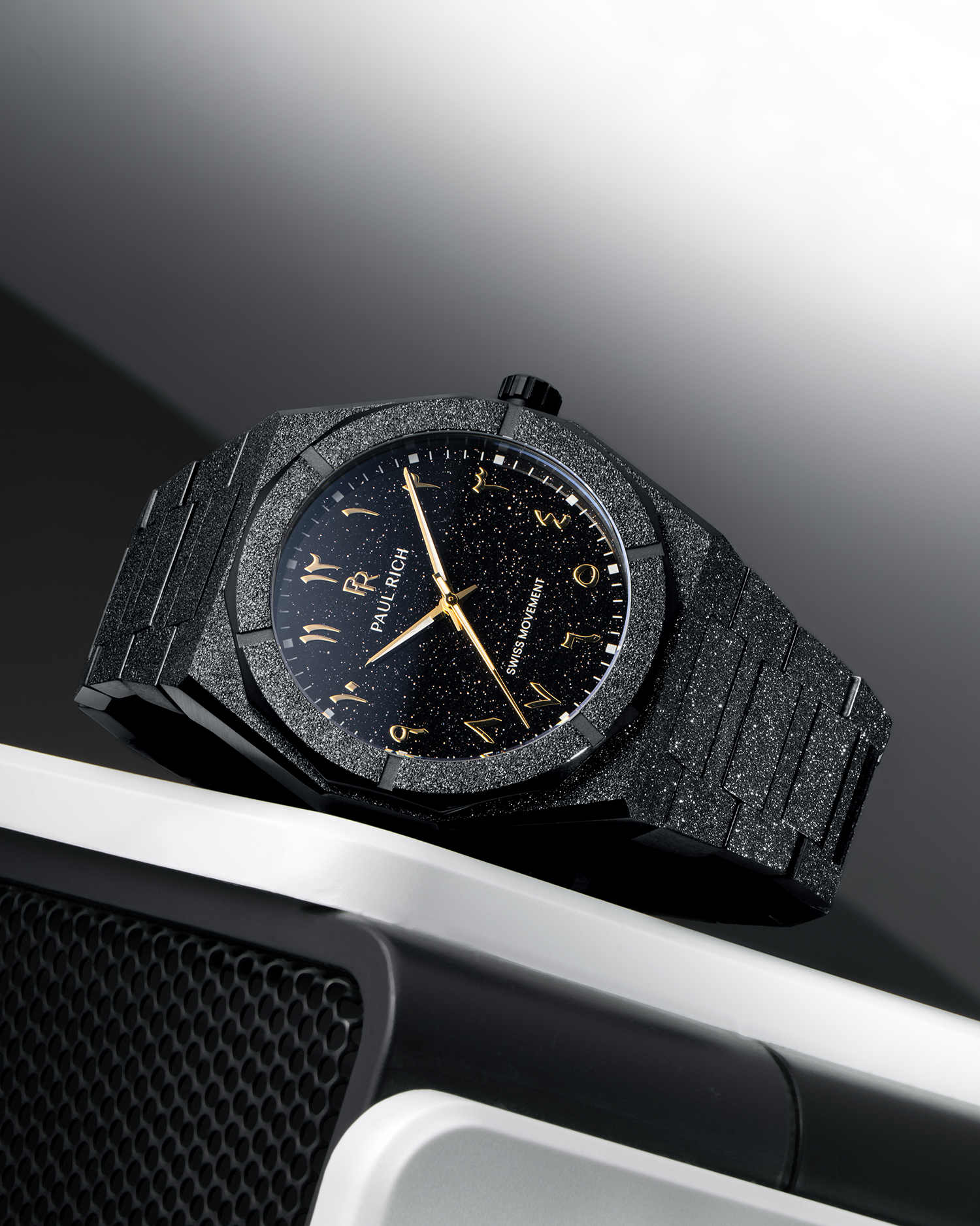 Paul Rich frosted black watch with black aventurine dial and gold Arabic numerals, resting on a silver and black speaker.
