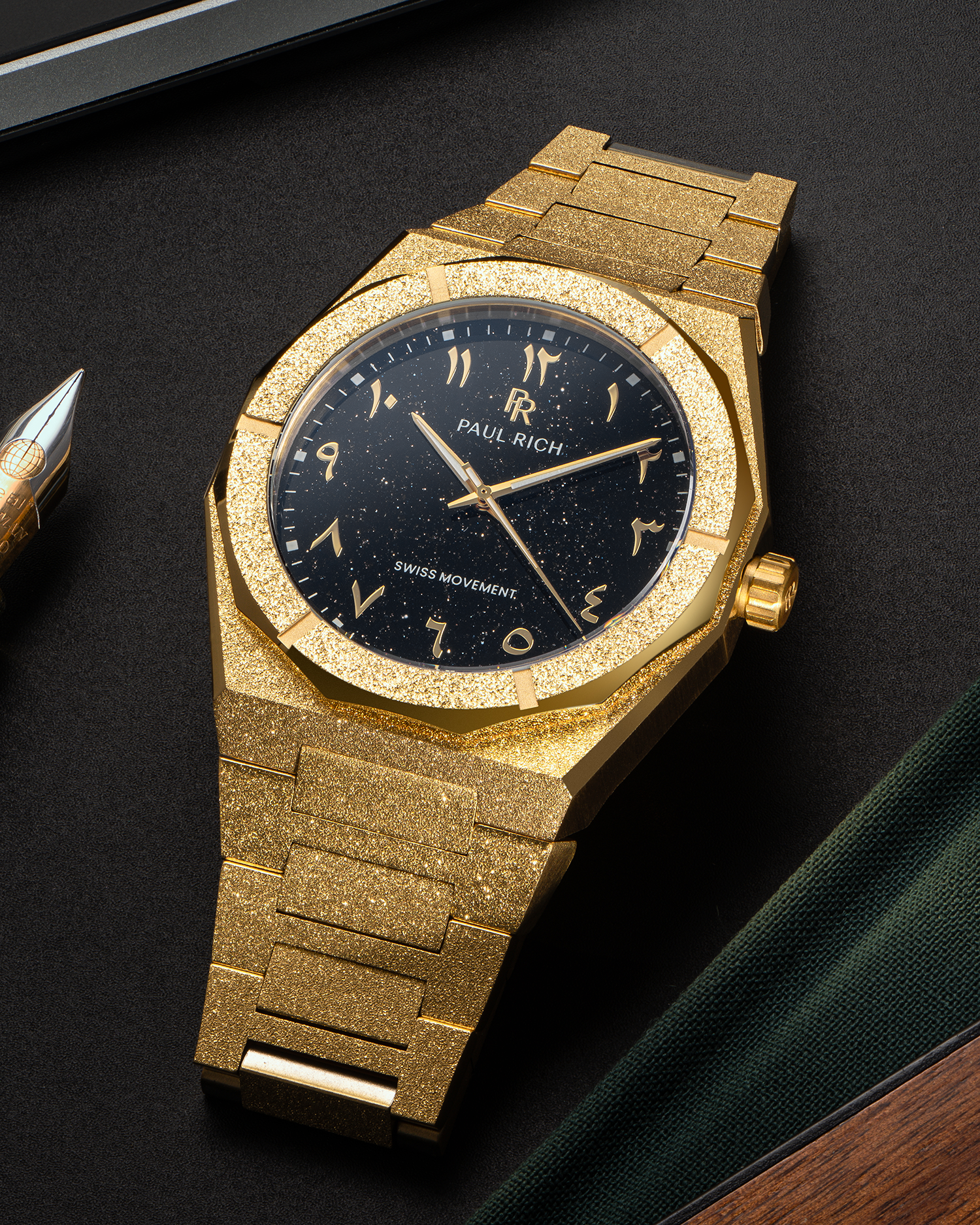 Paul Rich frosted gold watch with black aventurine dial and gold Arabic numerals, displayed on a black surface near a fountain pen.