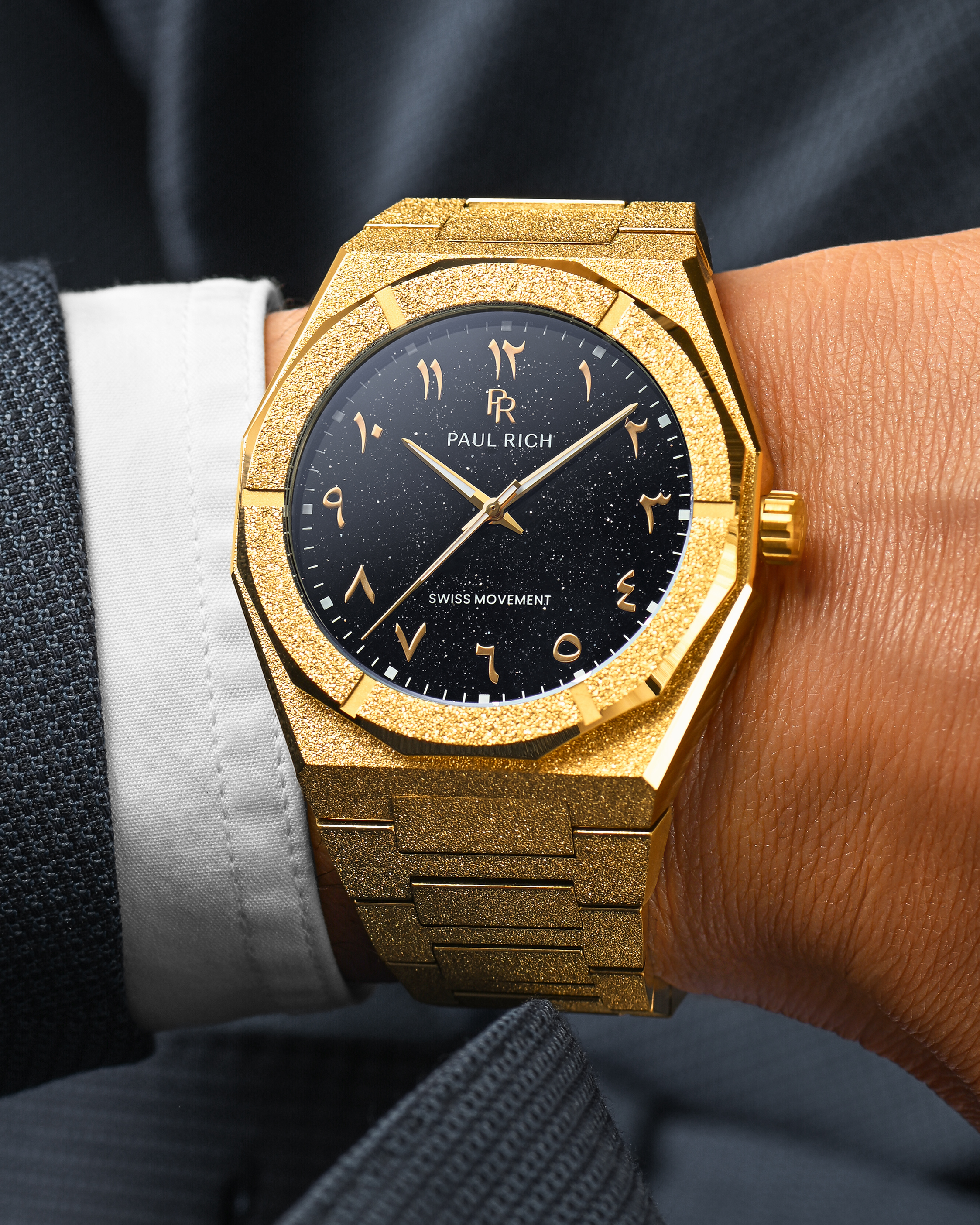 Close-up of Paul Rich frosted gold watch with Arabic numerals and black aventurine dial, worn on wrist with dark textured suit.