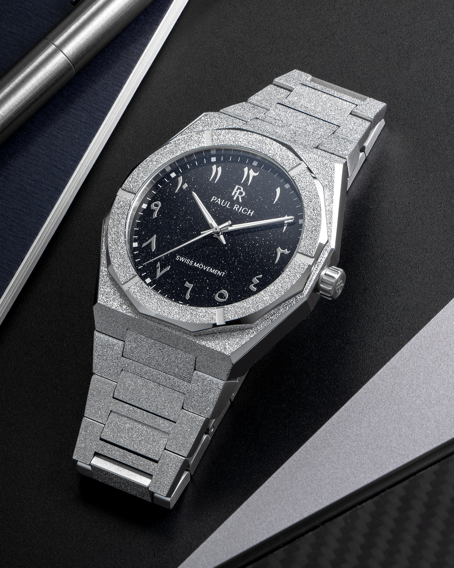 Paul Rich frosted silver watch with black aventurine dial and silver Arabic numerals, displayed on a black surface near a notebook.