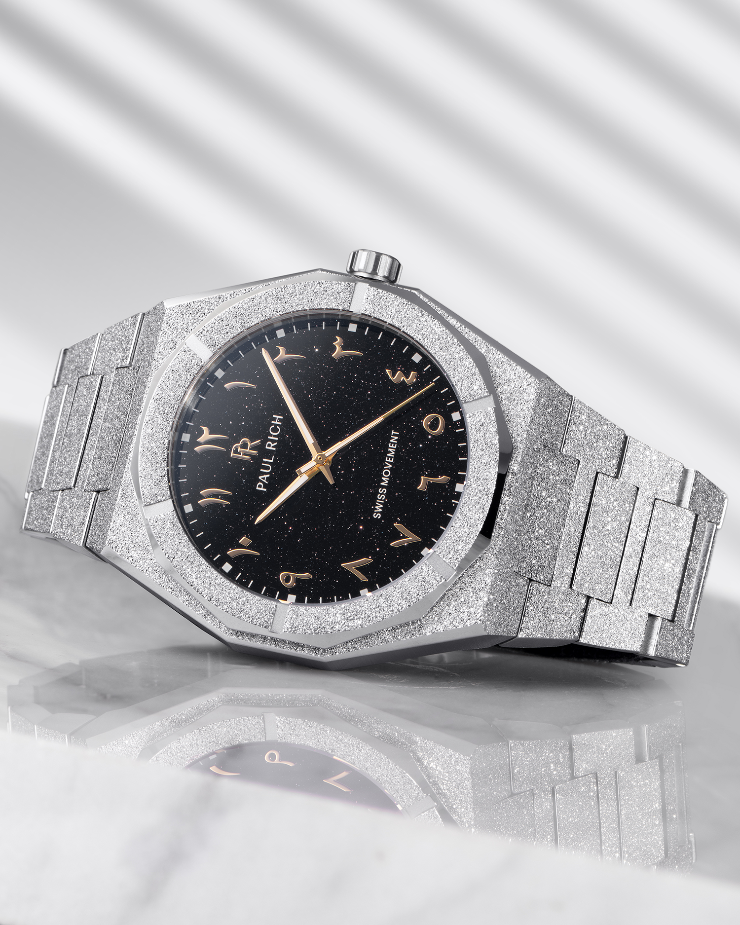 Paul Rich frosted silver watch with black aventurine dial and gold Arabic numerals, resting on a reflective marble surface.