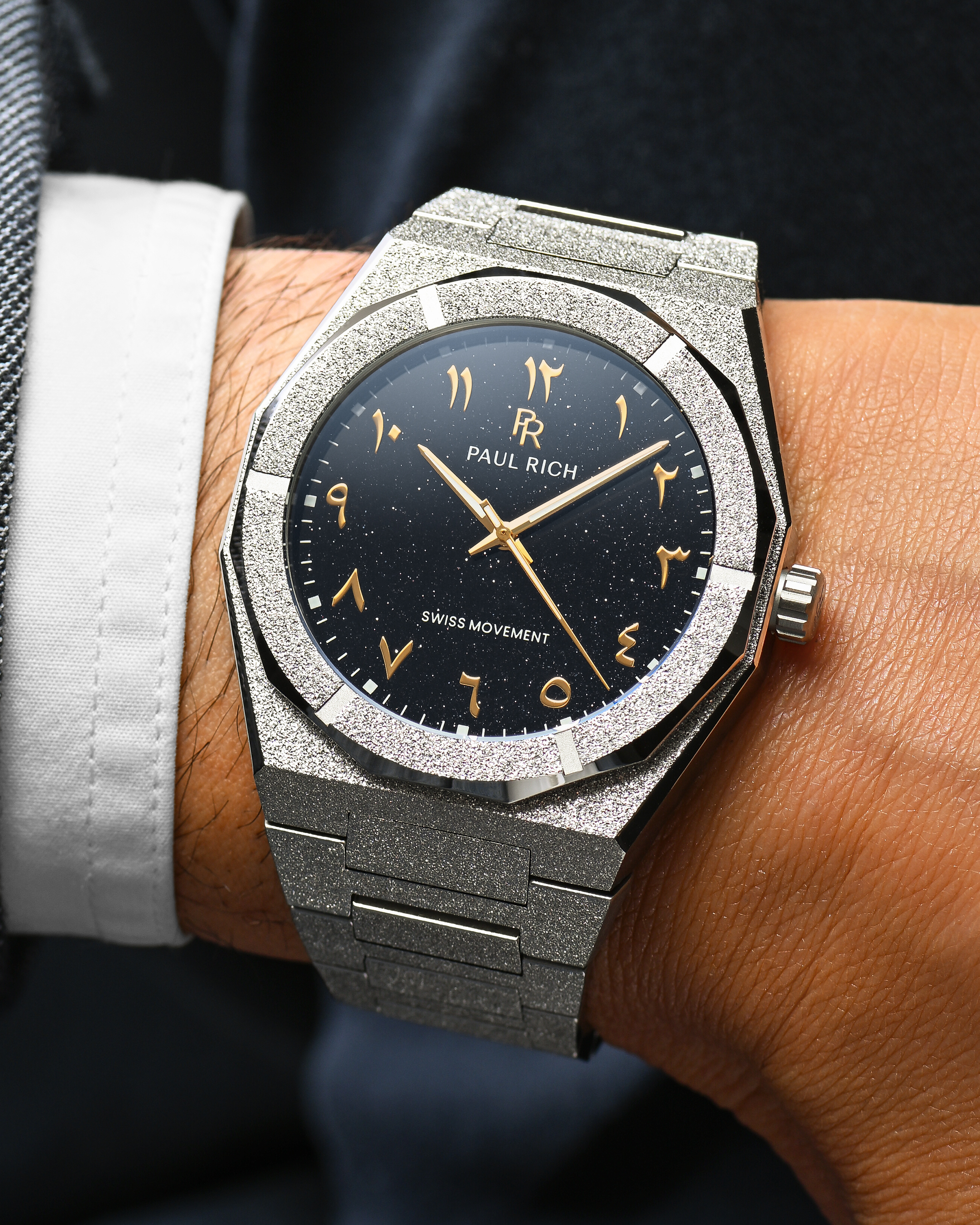 Close-up of Paul Rich frosted silver watch with gold Arabic numerals and black aventurine dial, worn with a white cuff and dark jacket.