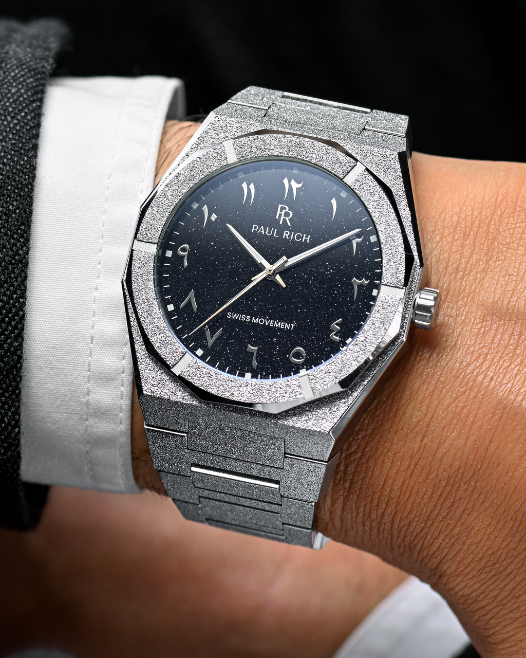Close-up of Paul Rich frosted silver watch with silver Arabic numerals and black aventurine dial, worn with white shirt and dark jacket.