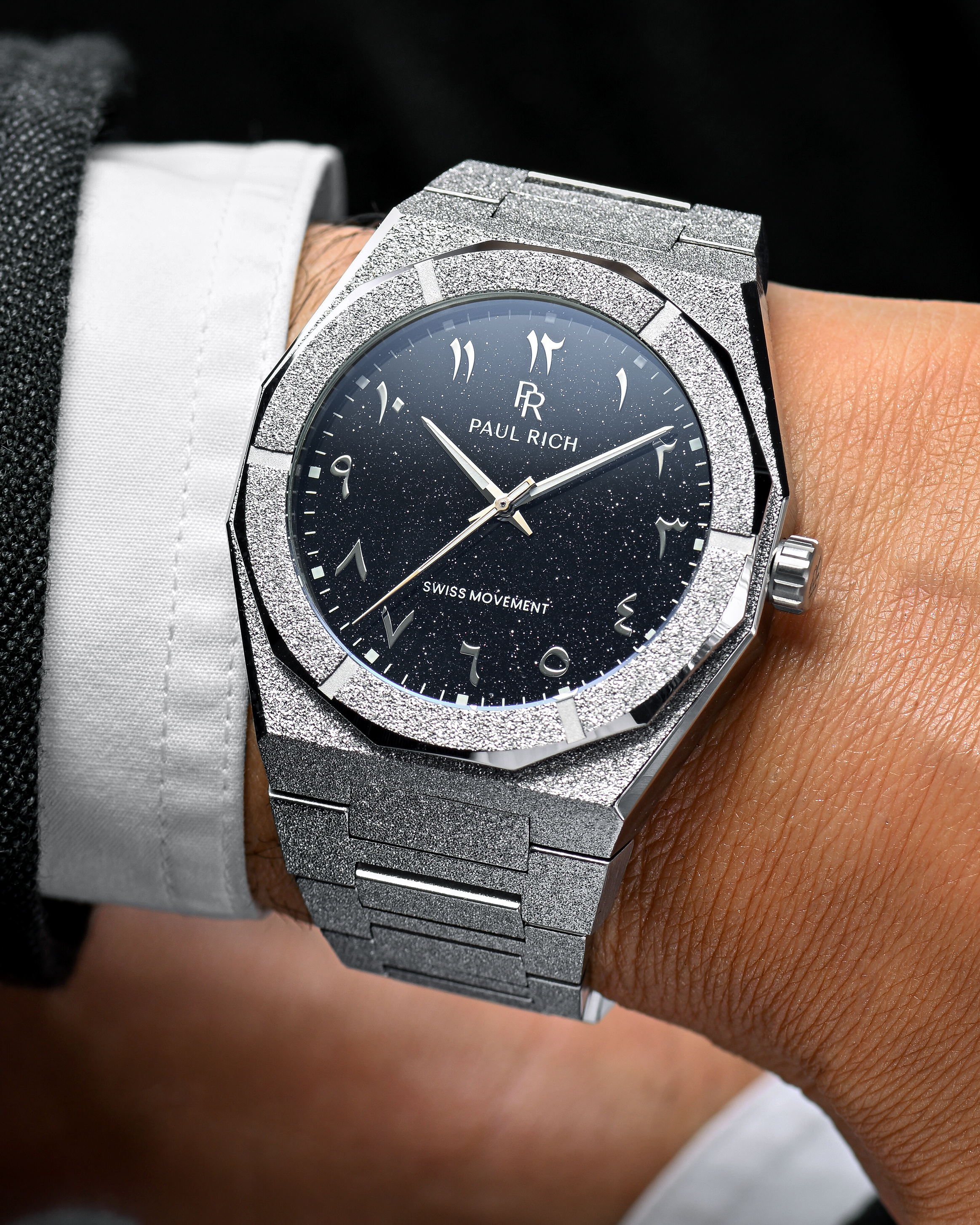 Close-up of Paul Rich frosted silver watch with silver Arabic numerals and black aventurine dial, worn with white shirt and dark jacket.