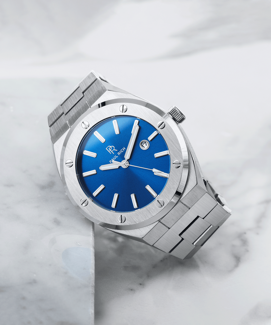 Paul Rich brushed silver watch with vivid blue sunray dial and date window, placed diagonally on white marble surface.