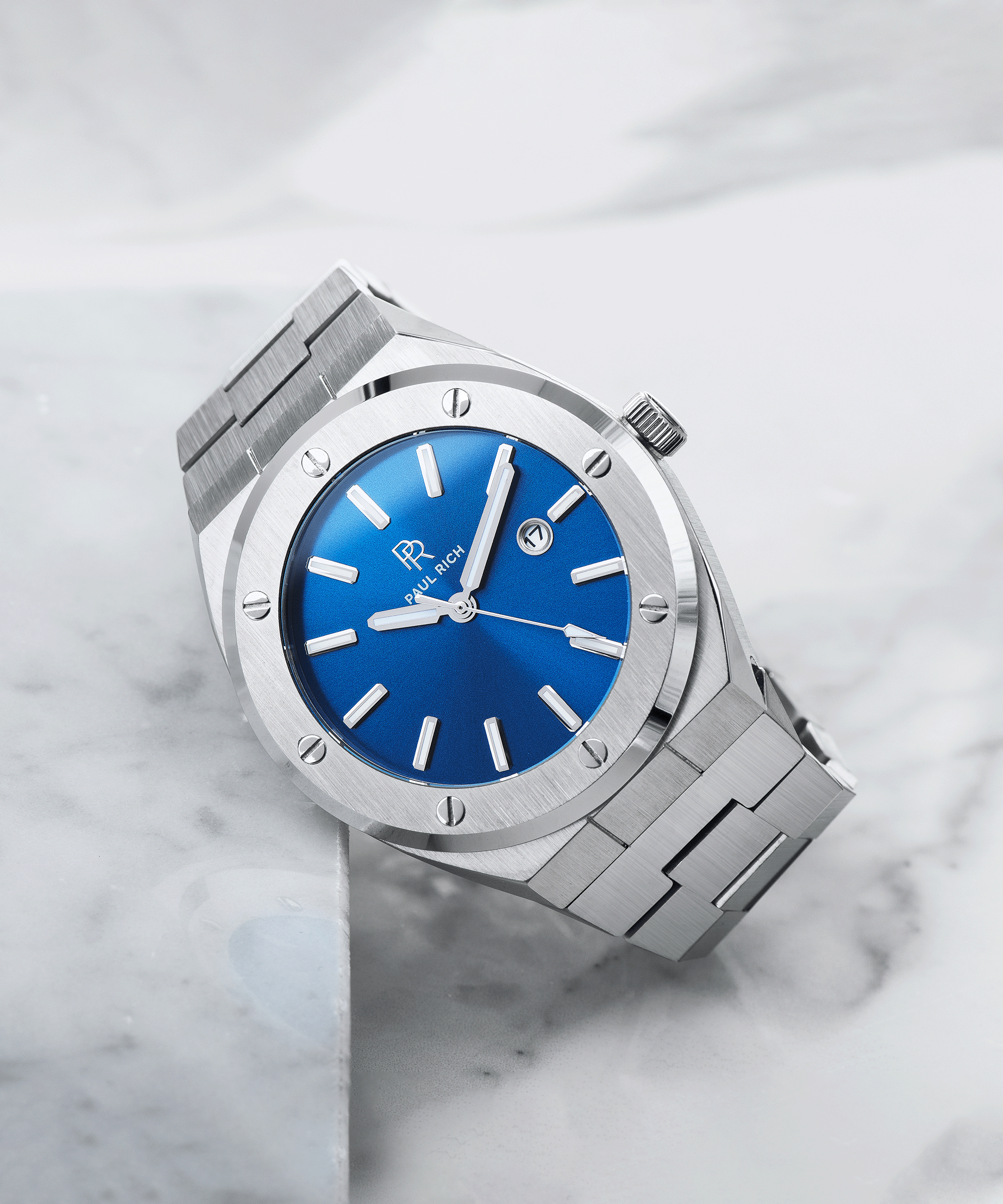 Paul Rich brushed silver watch with vivid blue sunray dial and date window, placed diagonally on white marble surface.